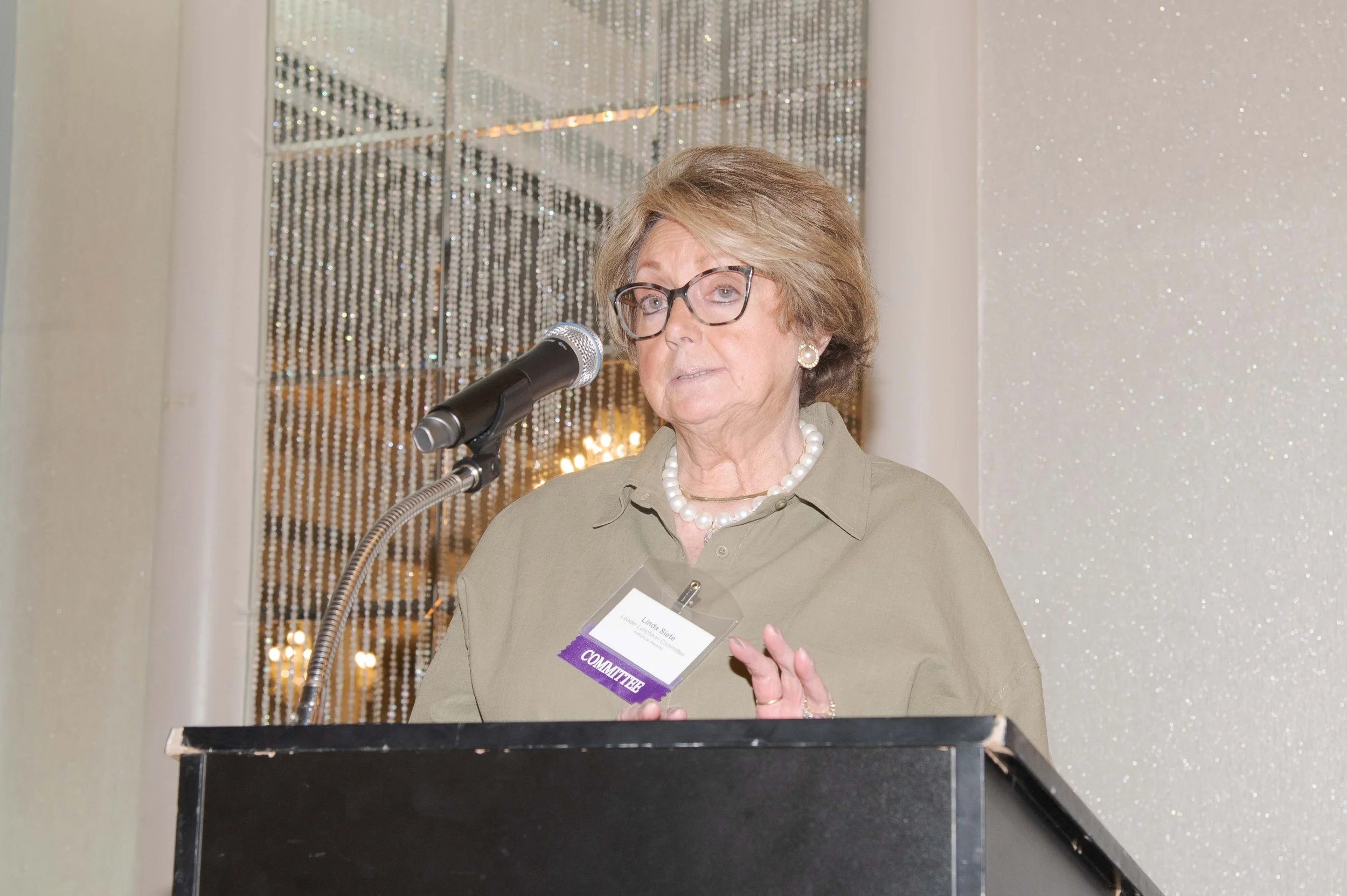 An older woman with glasses and earrings speaking at a podium, wearing a beige blazer with a name tag that reads 'Linda Siete' and a badge labeled 'COMMUNITY'. She is holding a piece of paper and standing in front of a sparkling backdrop.