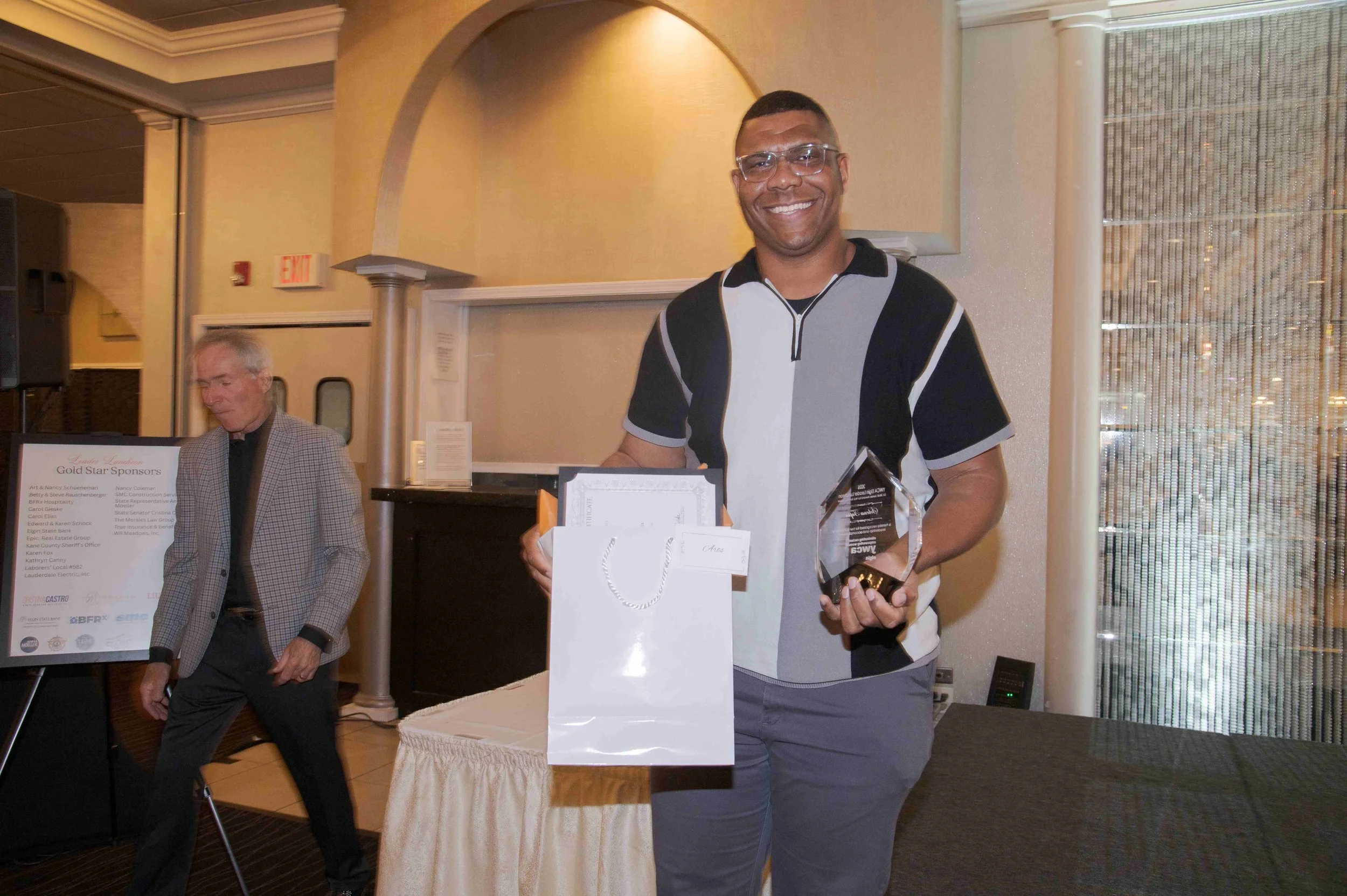 A man with glasses, smiling, holding an award, standing next to a table with partially visible awards and certificates, at an indoor event.