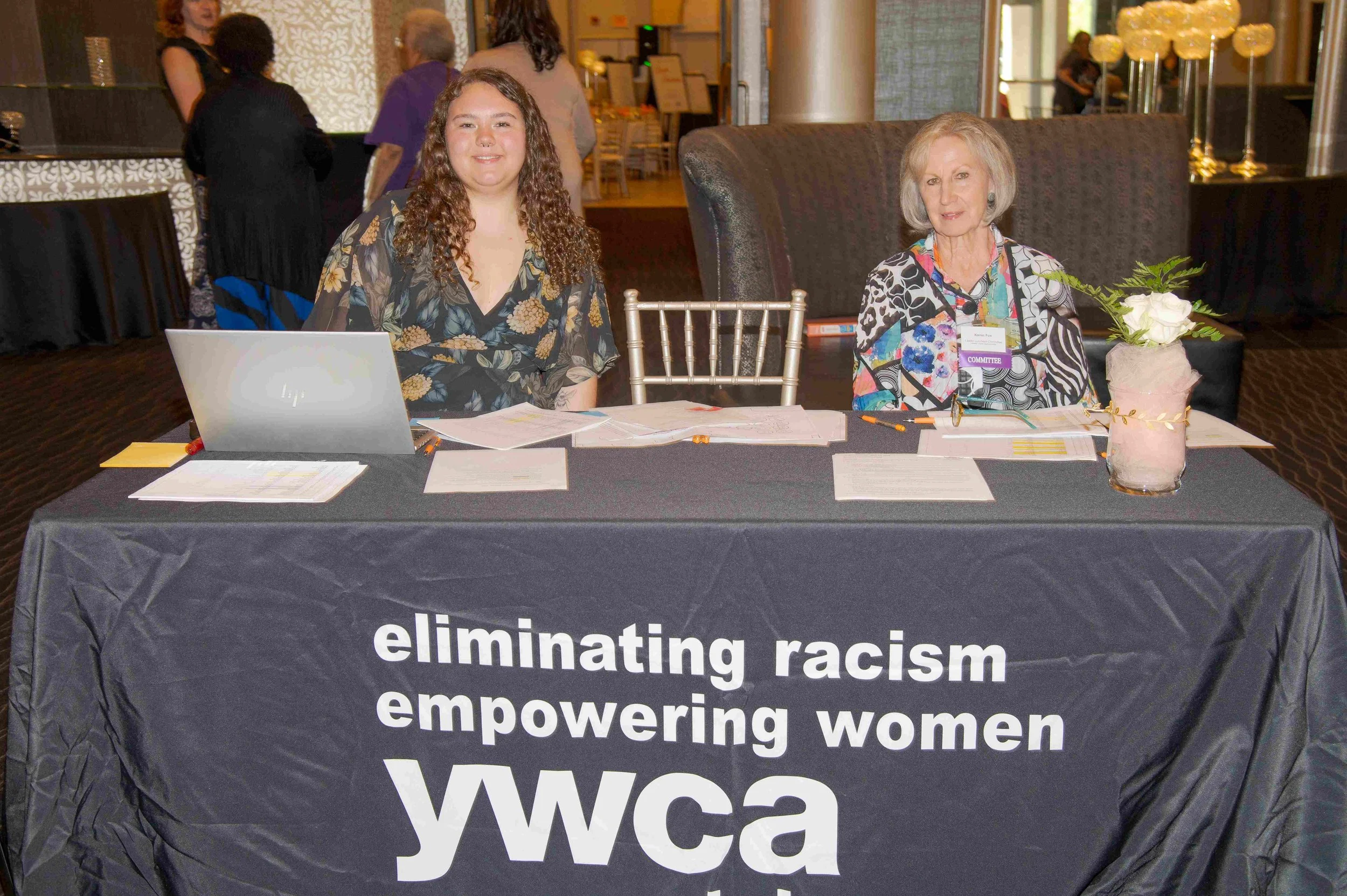 A woman sitting at a table with another young woman, in a setting that appears to be an event or conference. The table is covered with a black cloth featuring white text advocating against racism, promoting empowerment of women, and referencing the Y