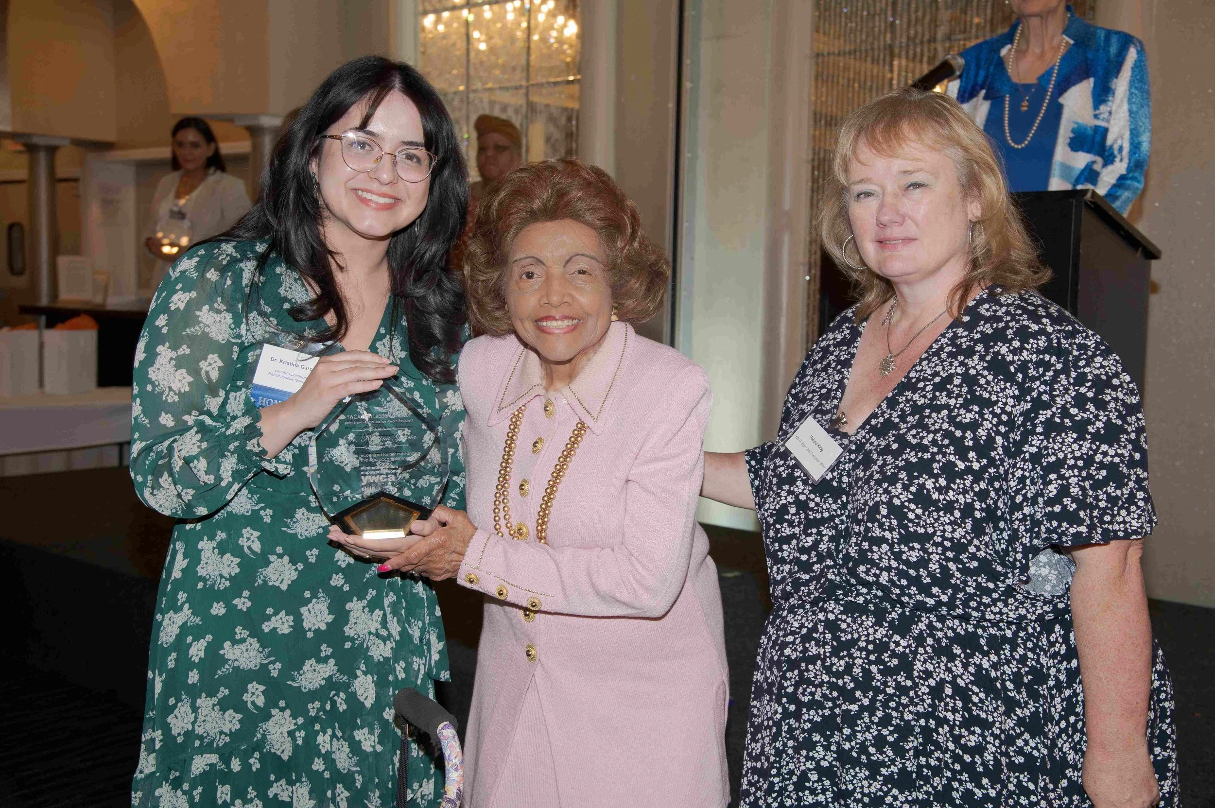 Three women smiling at an event, with one woman in the center holding an award. The woman on the left is wearing glasses and a green floral dress, the woman in the middle is wearing a pink outfit with pearl jewelry, and the woman on the right is wear