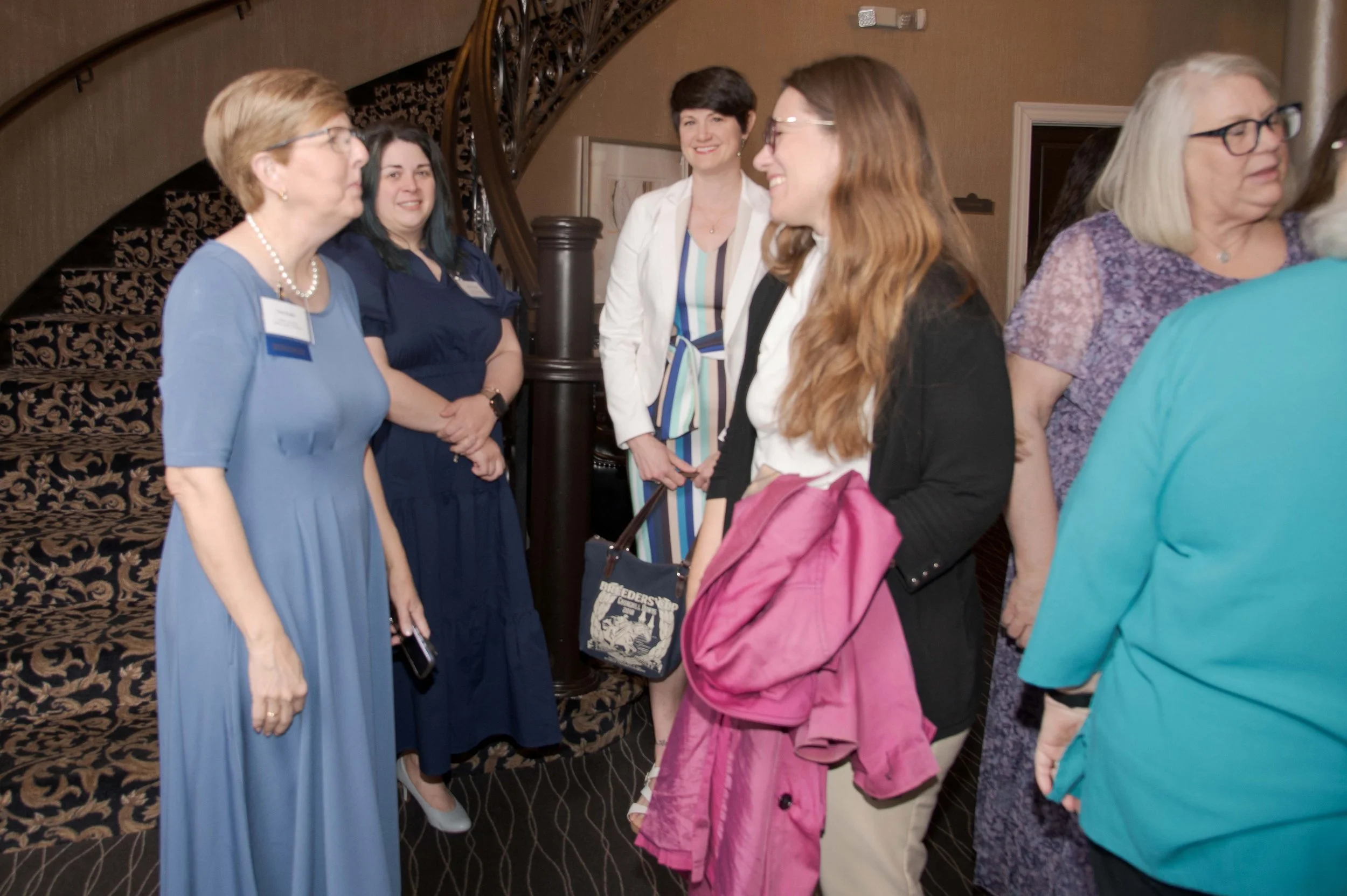 Group of women standing and talking in a hotel lobby with a staircase in the background.