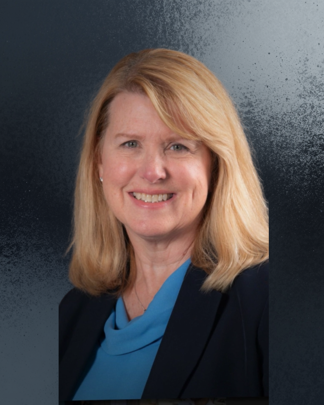 A smiling middle-aged woman with short brown hair, wearing a floral blazer, purple top, a necklace with a dark stone, and small earrings, against a blue mottled background.