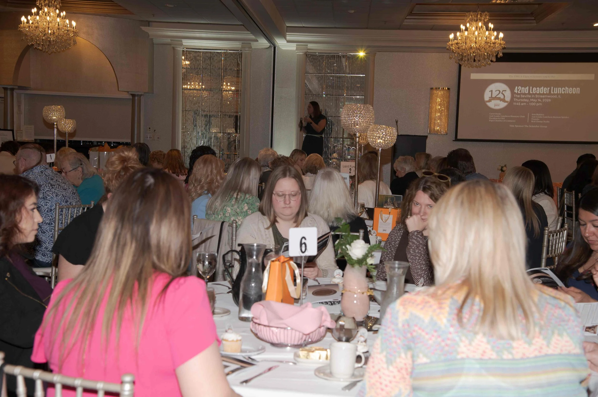 A large group of people sitting at round tables during a formal event in a decorated banquet hall, with a woman speaking on stage near a large screen displaying event details.