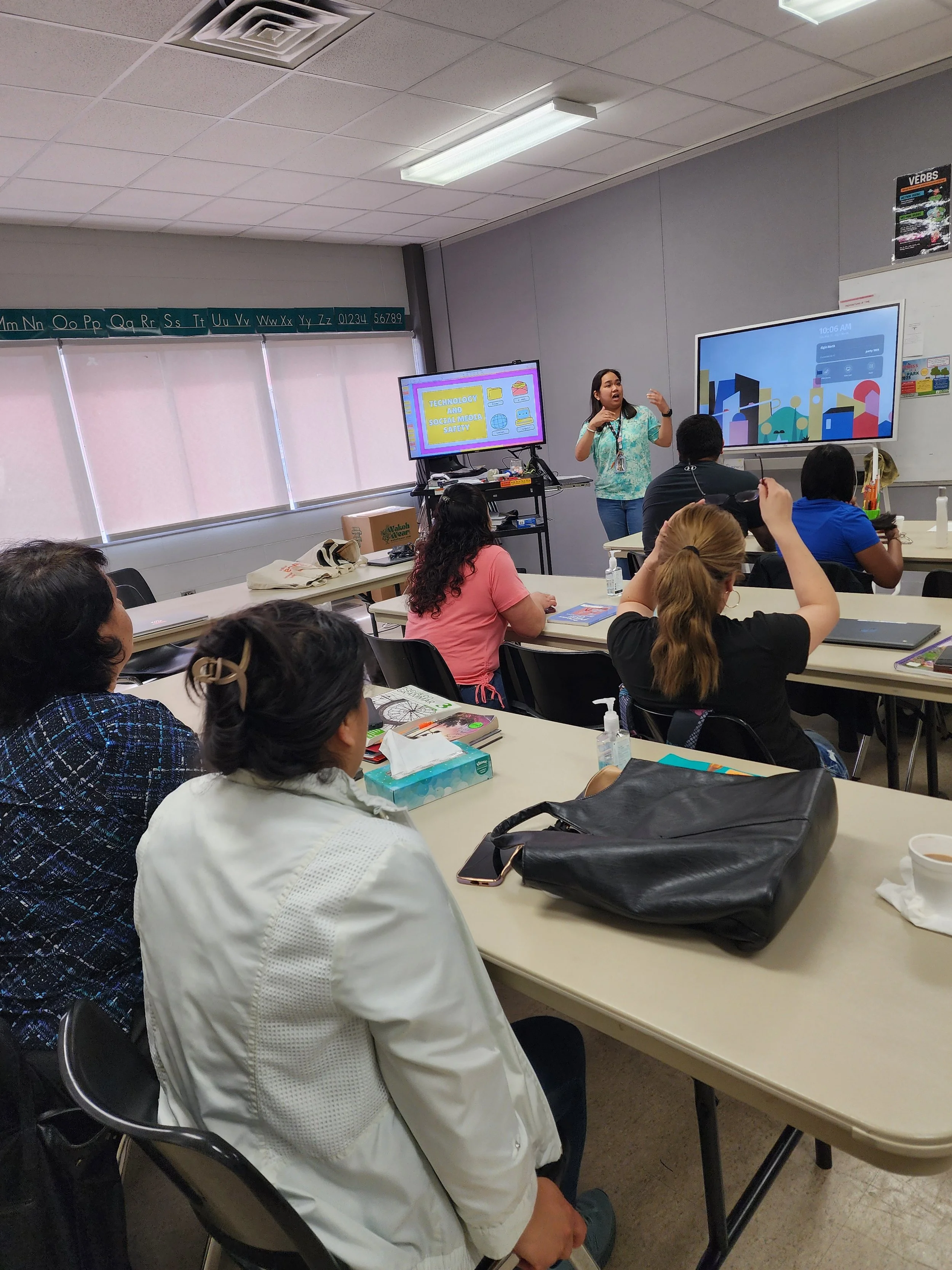 A classroom full of women sitting at tables, listening to a man in a checkered shirt speaking at the front. The women have notebooks, water bottles, and bags on the tables. A poster about face masks and a clock are on the wall.
