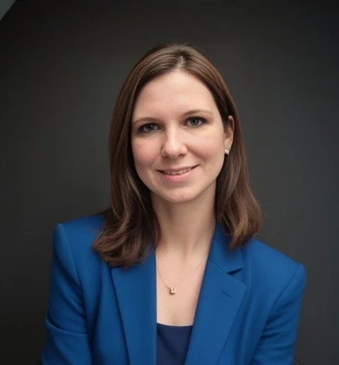 A woman with blonde, shoulder-length curly hair, wearing a white blazer over a light blue collared shirt, smiling at the camera against a plain gray background.