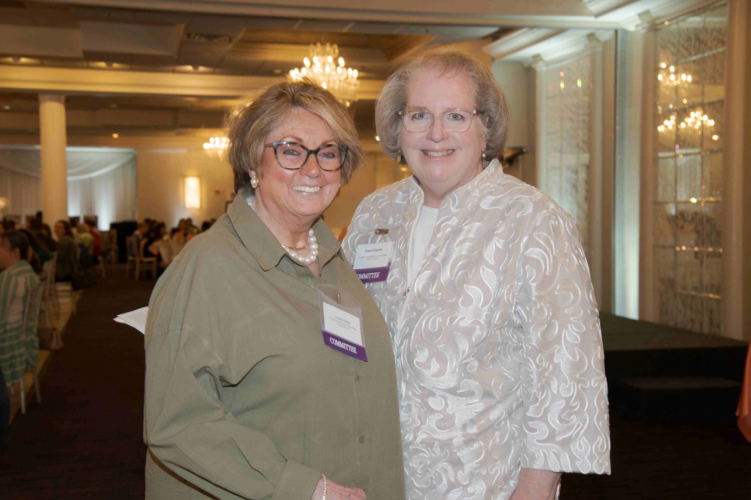 Two smiling women at a formal event, standing close together in a large, well-lit banquet hall with chandeliers and other guests seated at tables in the background.