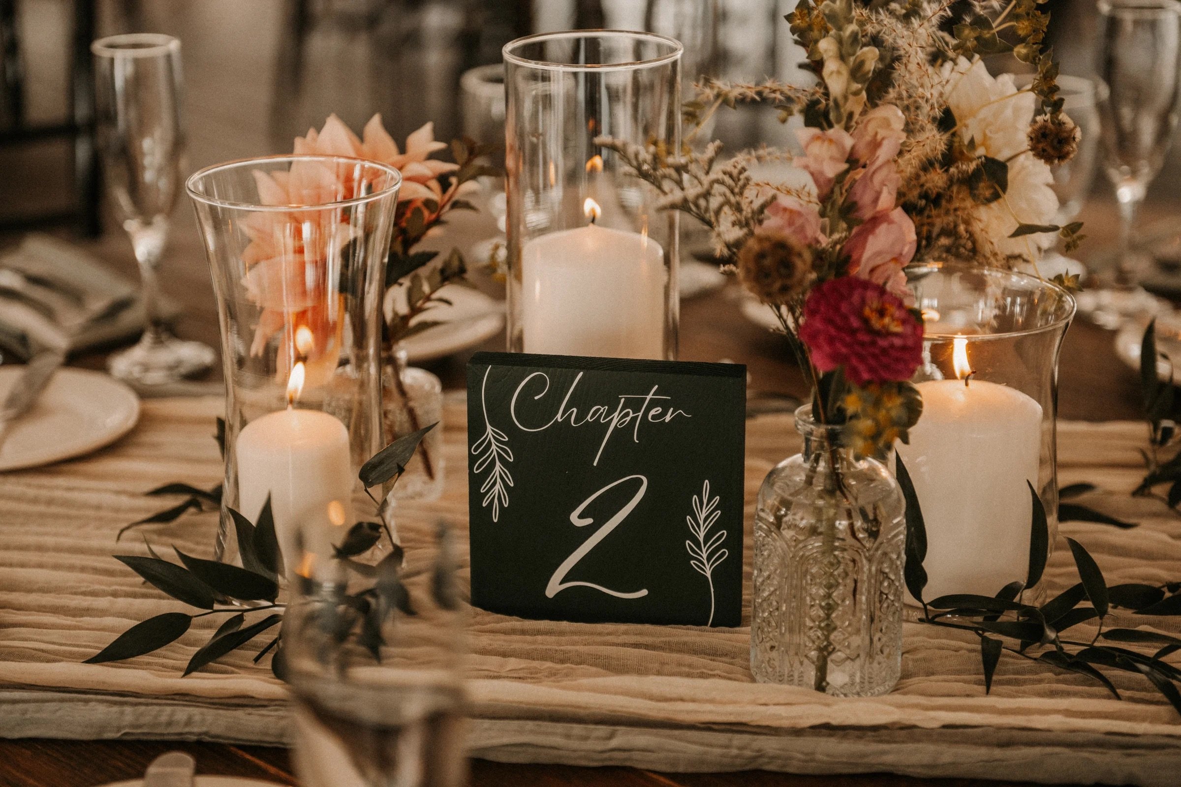 Table decorated with candles in glass holders, floral arrangements in glass vases, a black table number card reading 'Chapter 2', and a beige table runner.