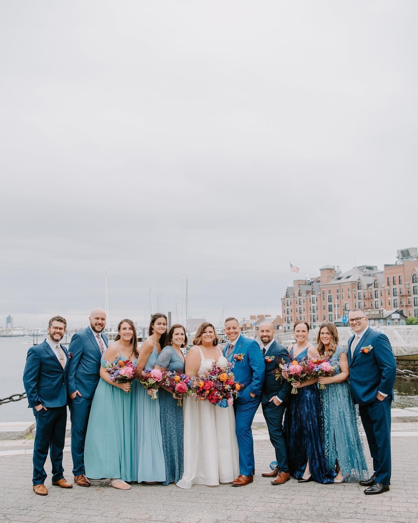 Group of brides and grooms in formal attire standing outdoors by water, with colorful bouquets, smiling for a wedding photo.