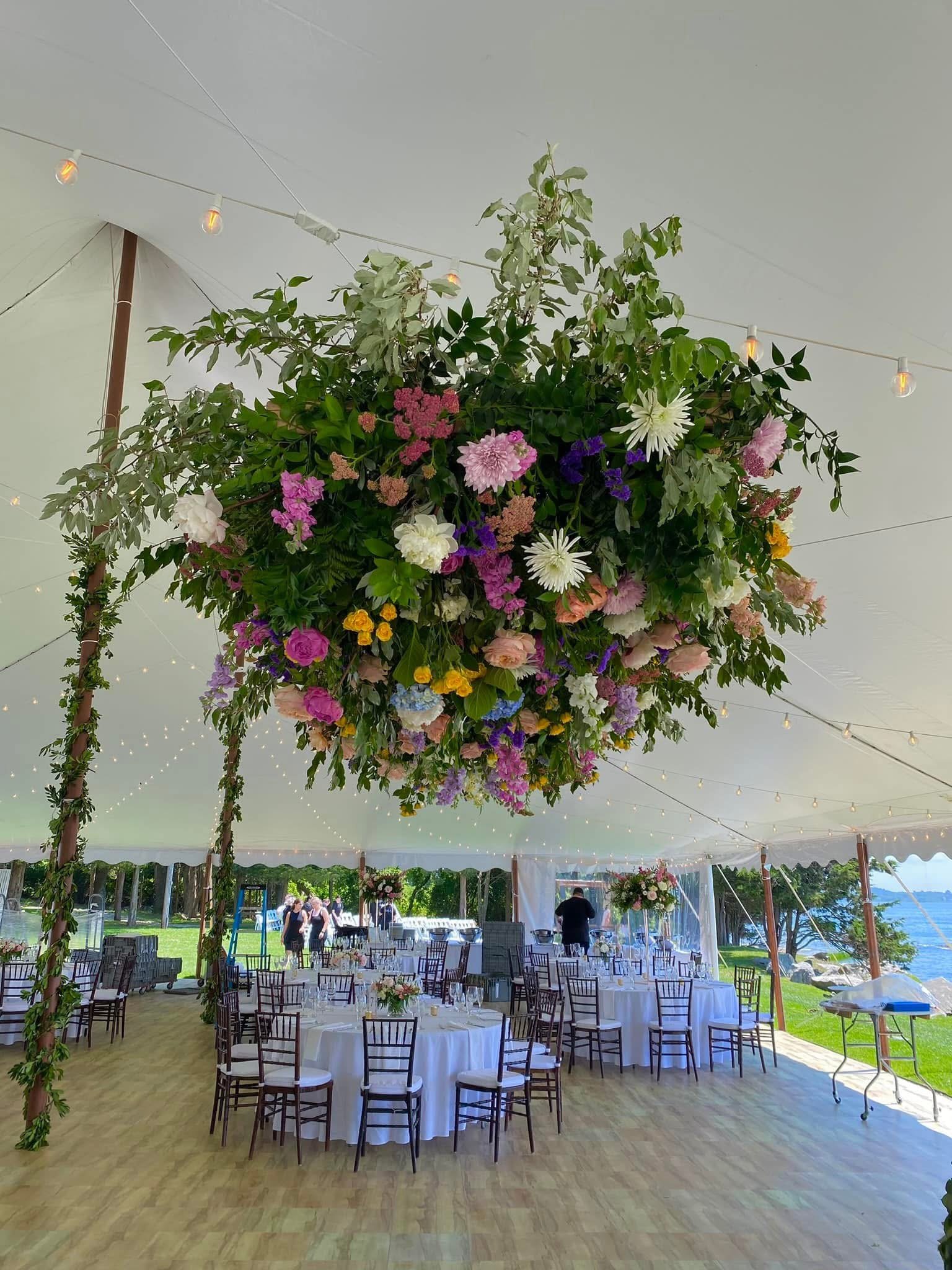 A large floral arrangement suspended from a tent ceiling at a wedding reception, with tables and chairs set for guests near a lakeside.