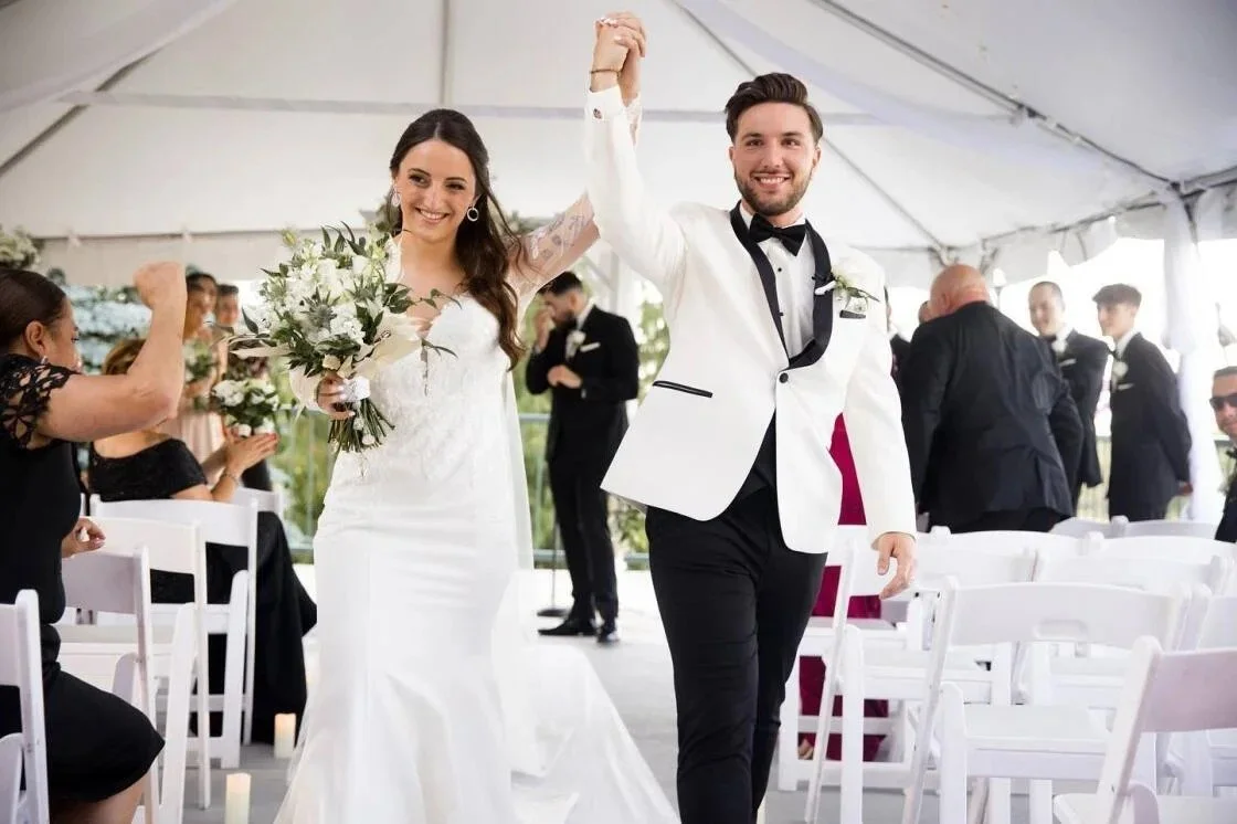 A newlywed couple walking down the aisle in a wedding tent, holding hands with the bride holding a bouquet of white flowers, surrounded by seated and standing guests in formal attire.