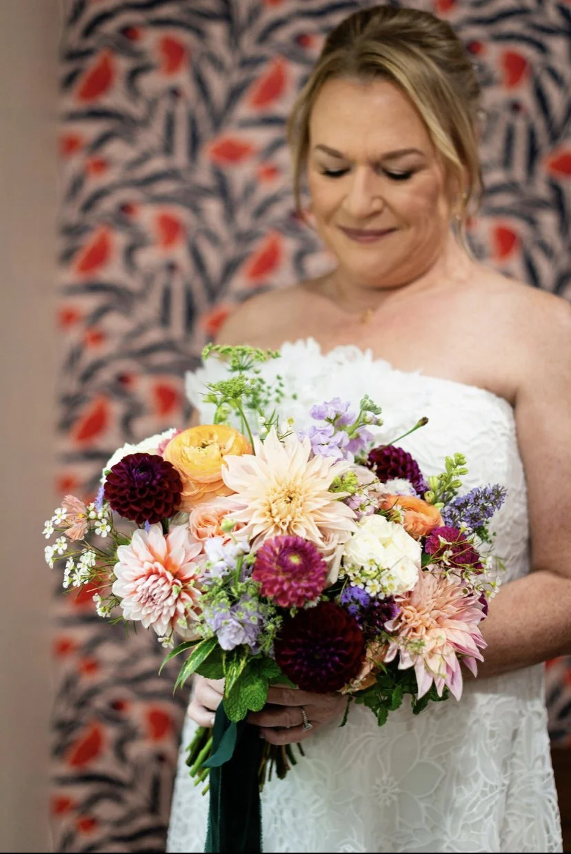 A woman in a white dress holding a colorful bouquet of flowers, standing in front of a patterned background.