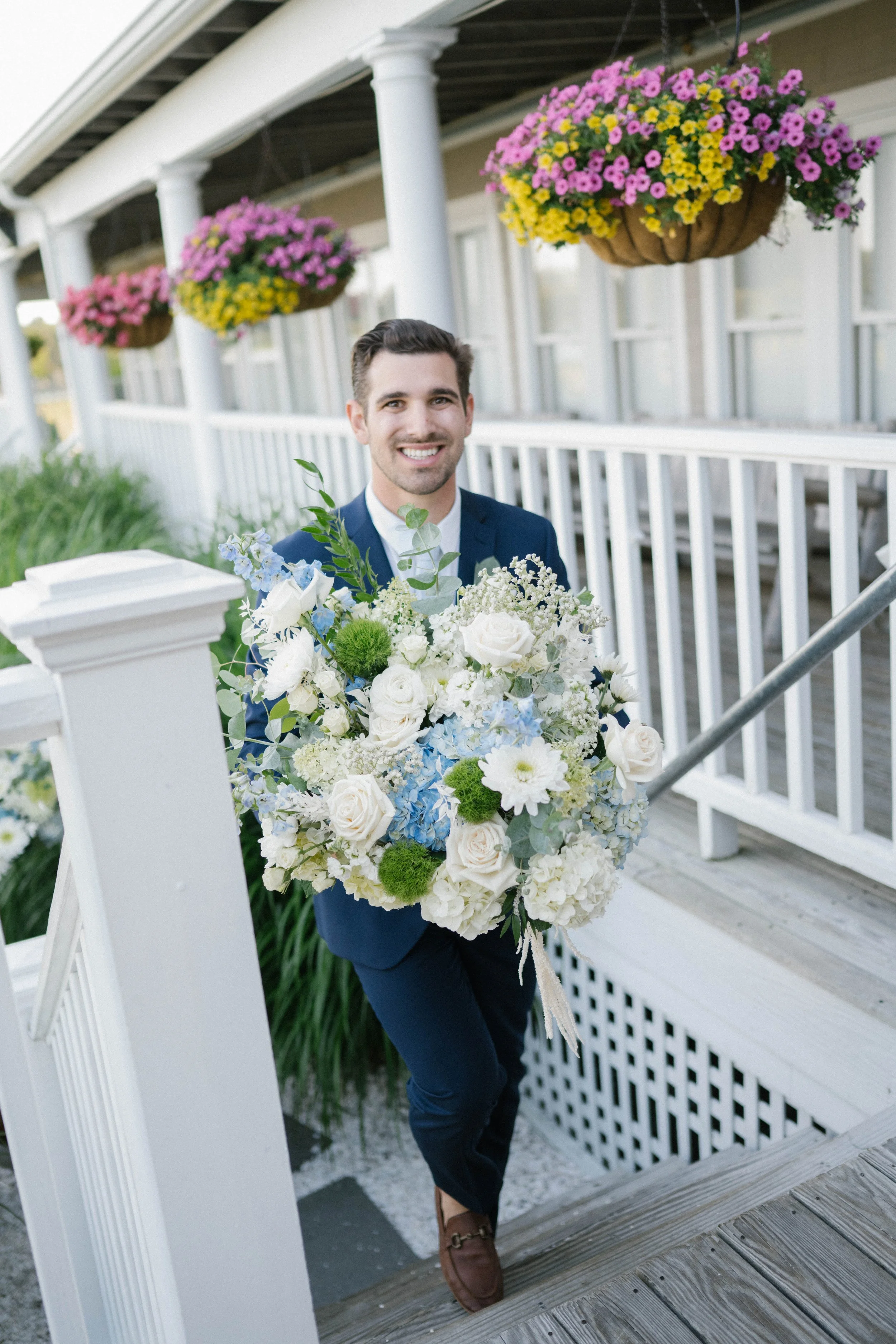 Man in a blue suit holding a large floral bouquet, smiling on a porch with hanging pink and yellow flowers.