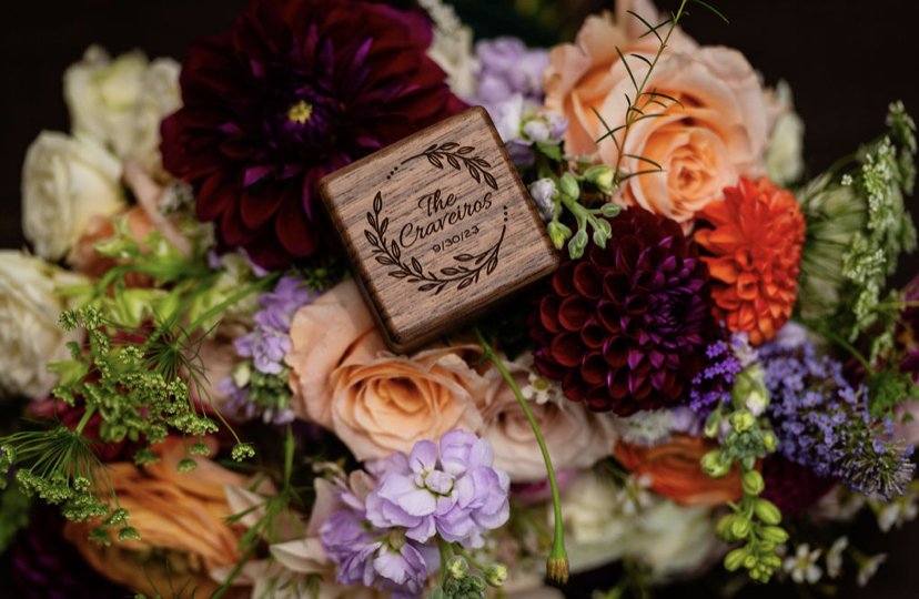 A floral arrangement with a small wooden box engraved with 'The Cravenos 01.30.23' placed among various colorful flowers including roses, dahlias, and other blossoms.