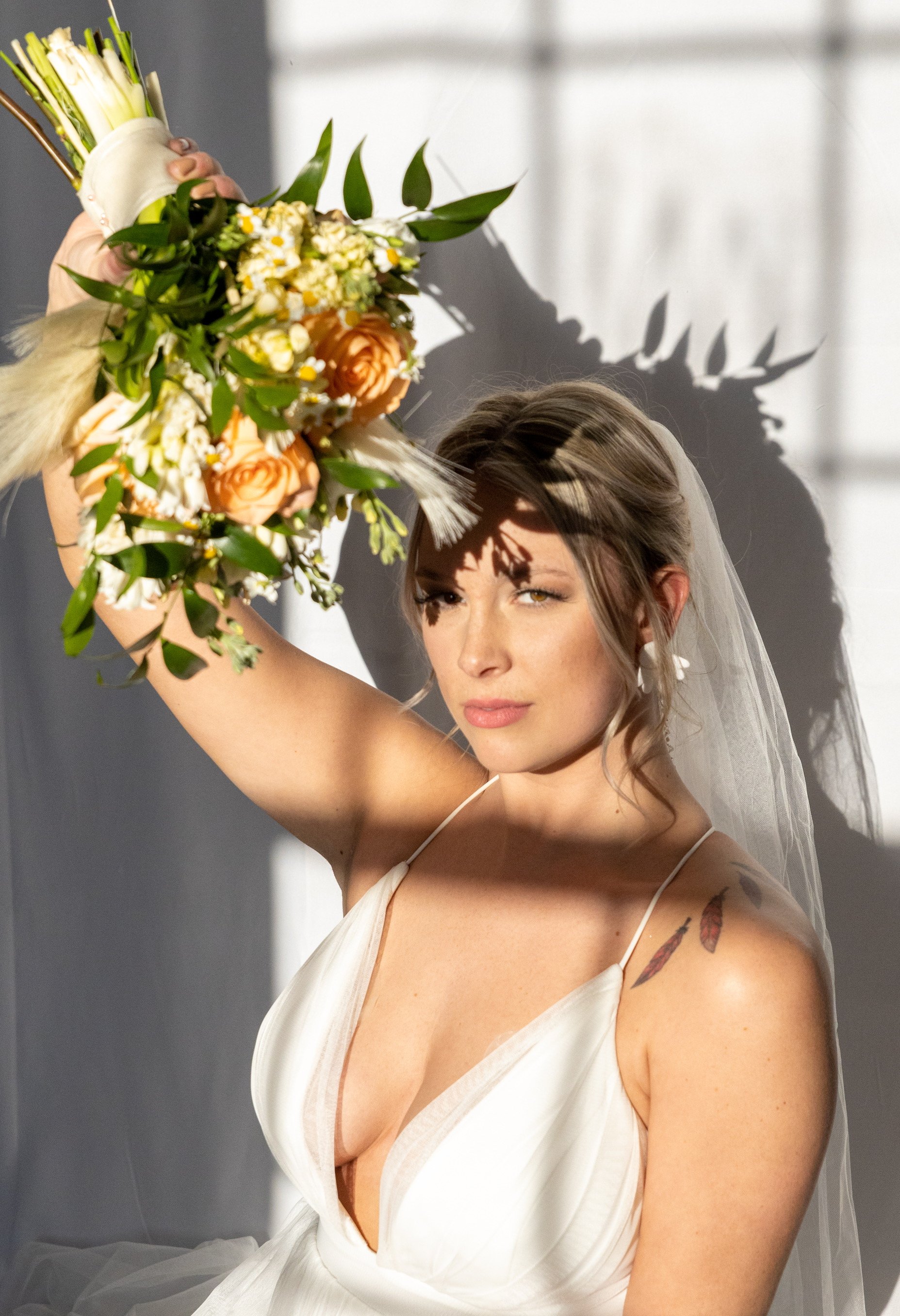 Bride in a white wedding dress with a veil holds a bouquet of flowers, casting a shadow of leaves on her face, sunlight illuminating her