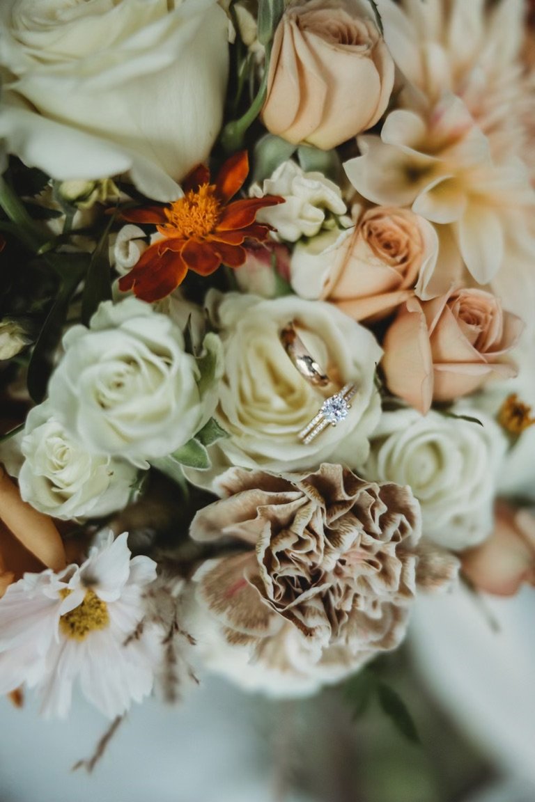 Close-up of a wedding bouquet with white, peach, and cream-colored roses, some dried flowers, and a diamond engagement ring nestled among the flowers.