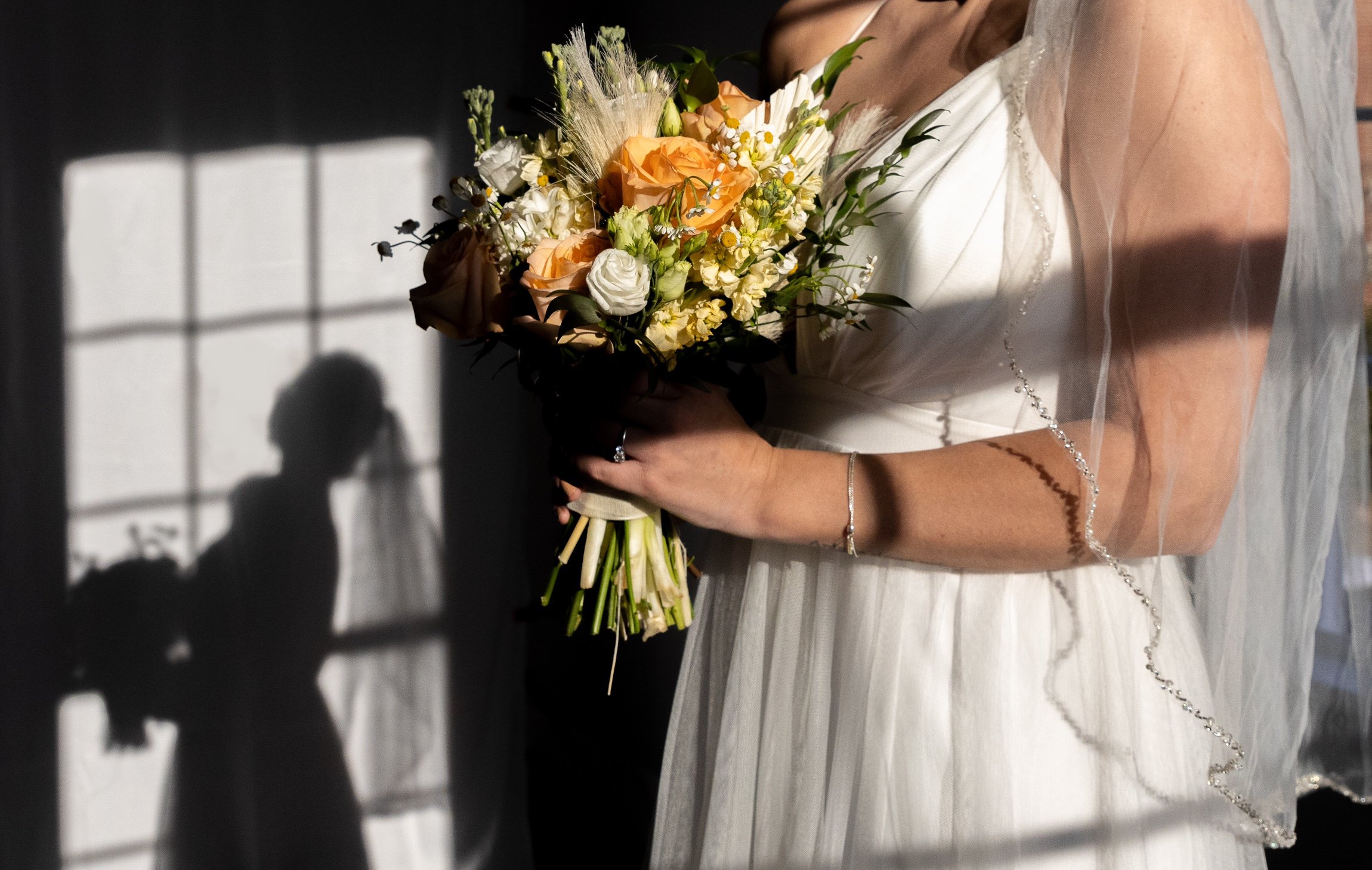 A bride holding a bouquet of peach, white, and green flowers with her shadow cast on a wall by sunlight through a window.