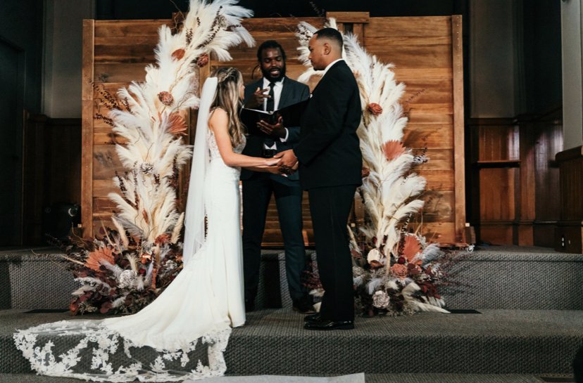 A couple getting married at a ceremony with an officiant, holding hands and looking at each other, against a decorated wooden backdrop with pampas grass and flowers.