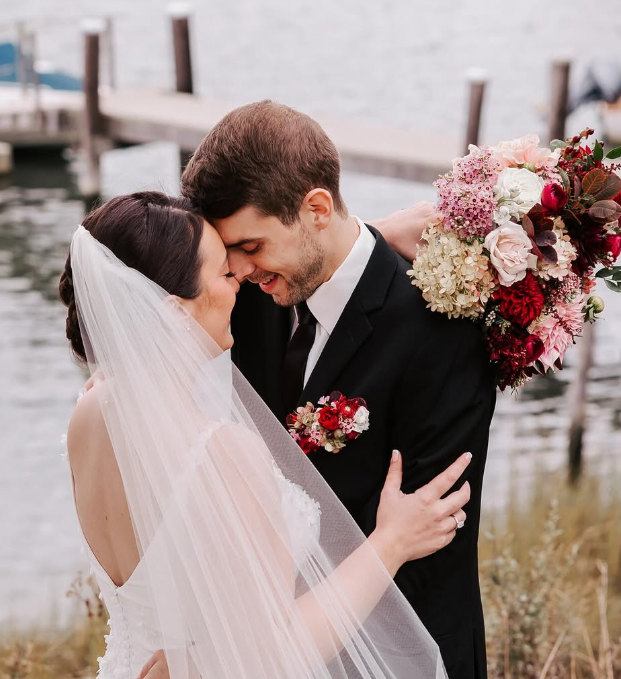 Bride and groom sharing a close moment by the water, with the groom holding a bouquet of pink, white, and red flowers.