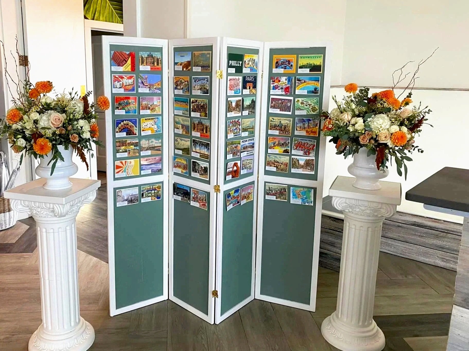 A photo of a display board with various colorful postcards or pictures, flanked by two white columns each topped with floral arrangements, inside a room with a wooden and tiled floor.