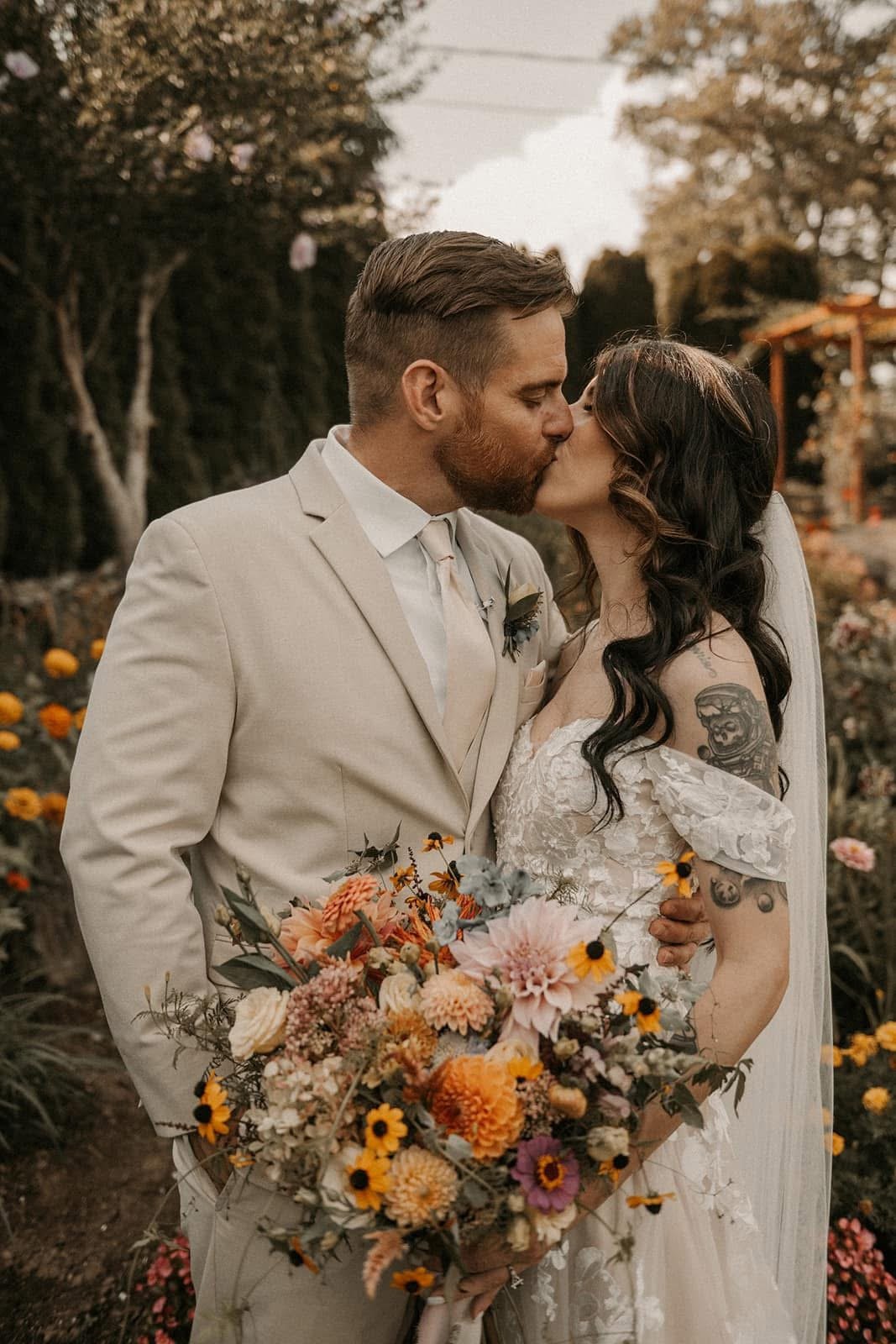 Bride and groom kissing outdoors during wedding, with the bride holding a large colorful bouquet, in a garden with flowers and trees in the background.