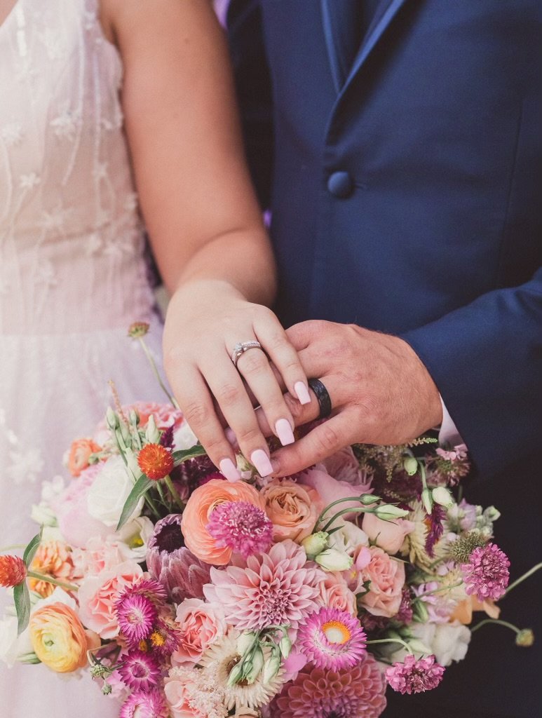 Close-up of a bride and groom holding hands over a bouquet of pink and peach flowers, showcasing wedding rings on the bride's finger and a dark wedding band on the groom's finger.