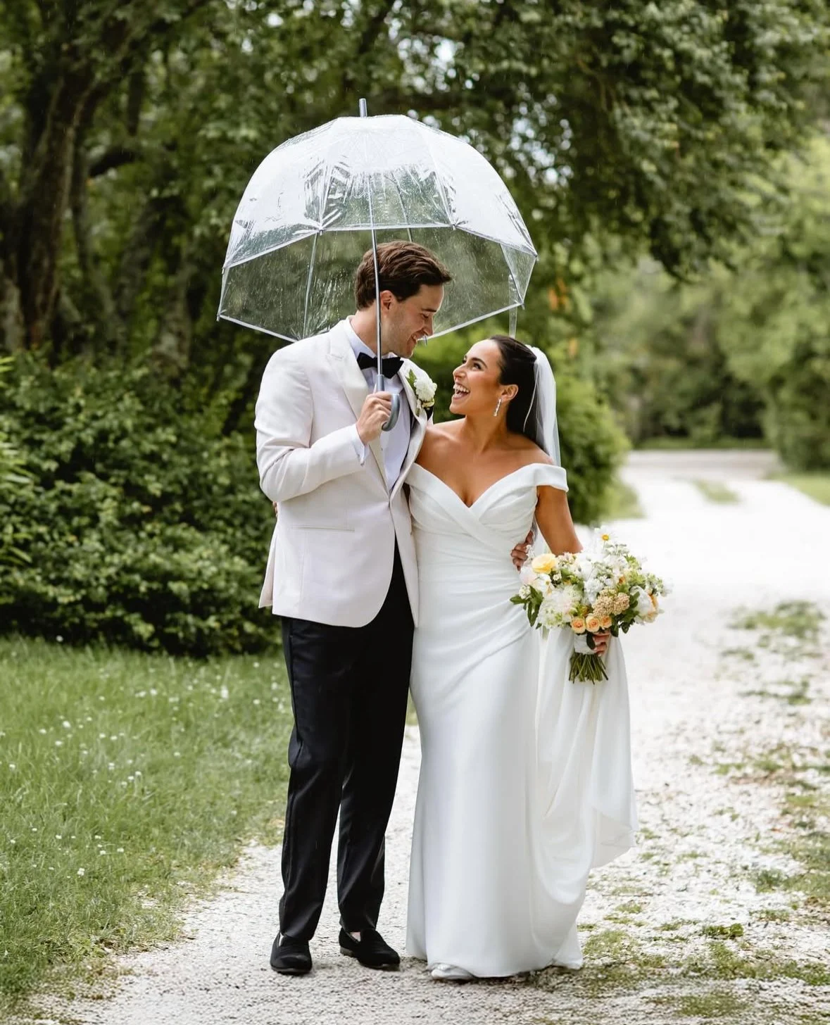 A bride and groom standing under a clear umbrella outdoors, smiling at each other on a rainy day. The bride is holding a bouquet of flowers and wearing a white wedding dress, while the groom is dressed in a white tuxedo jacket and black pants.