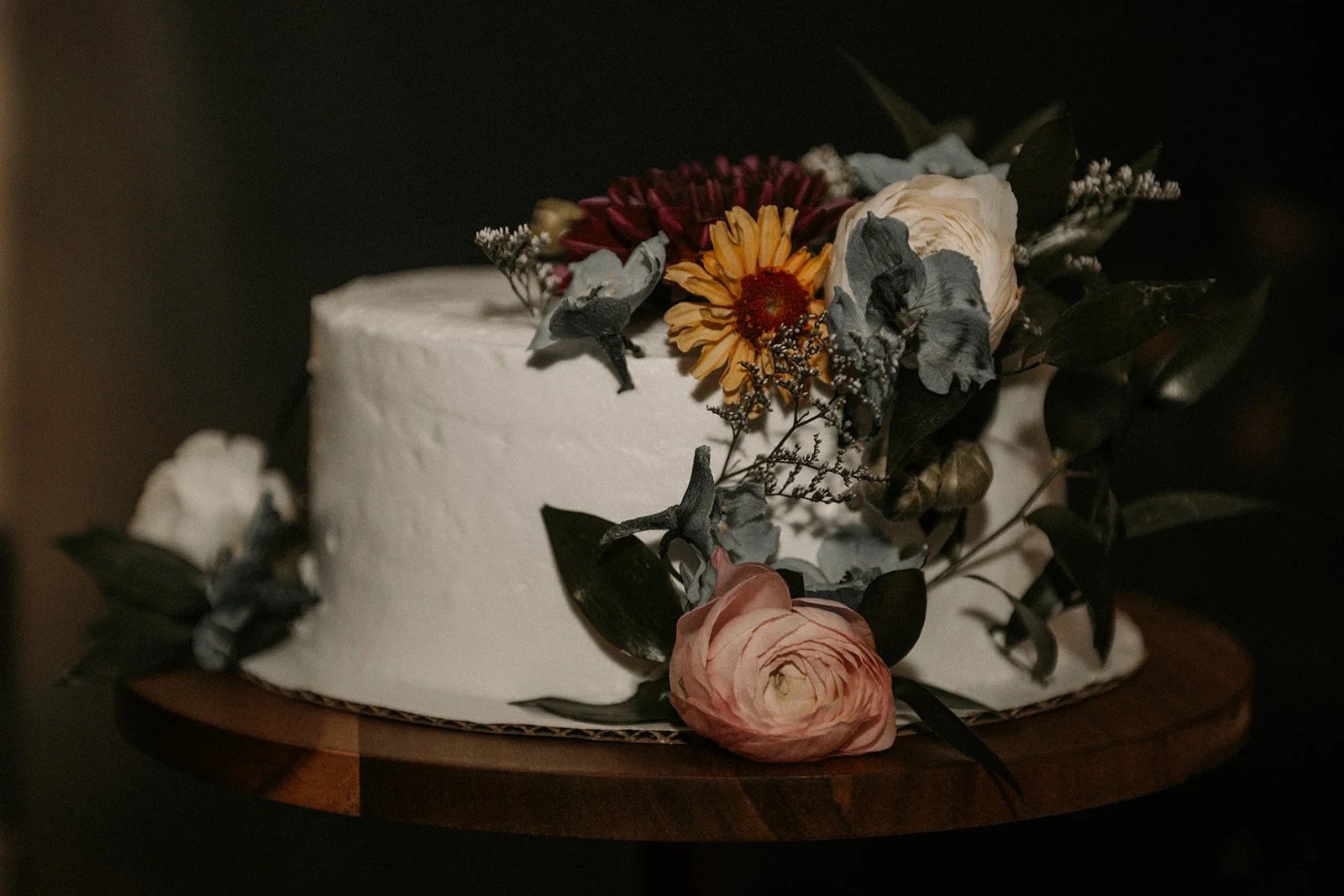 A white frosted cake decorated with a variety of dried flowers including roses and greenery, sitting on a round wooden cake stand.