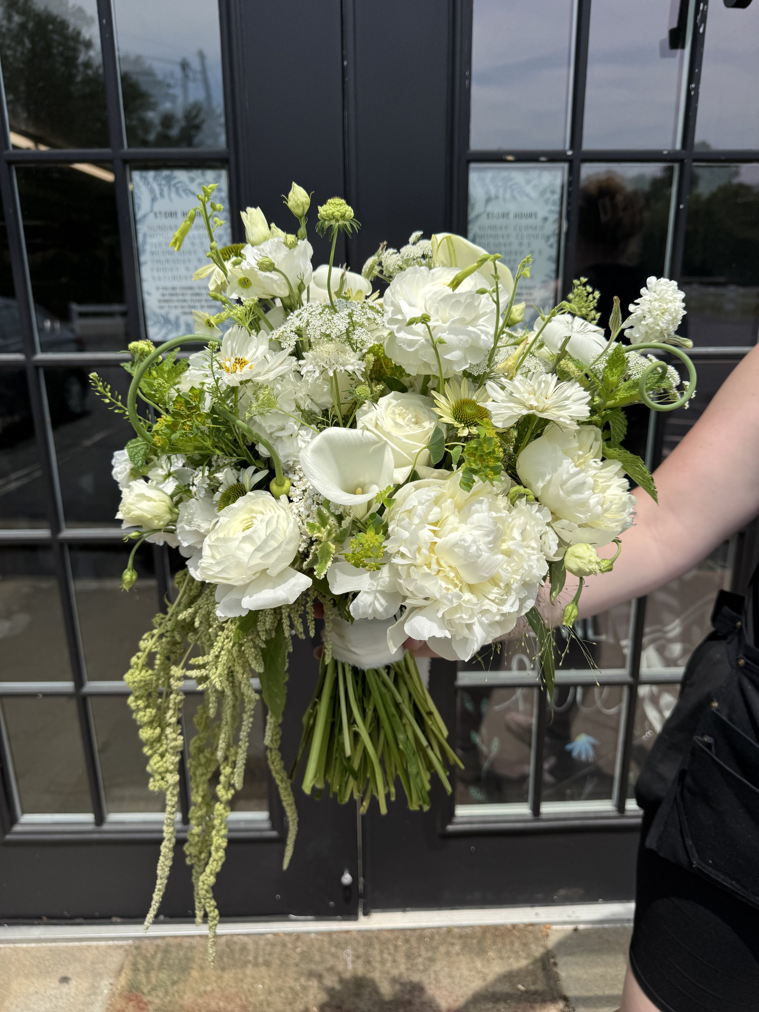 Person holding a large bouquet of white flowers, including roses, peonies, and other white blooms, in front of a black door with glass panes.