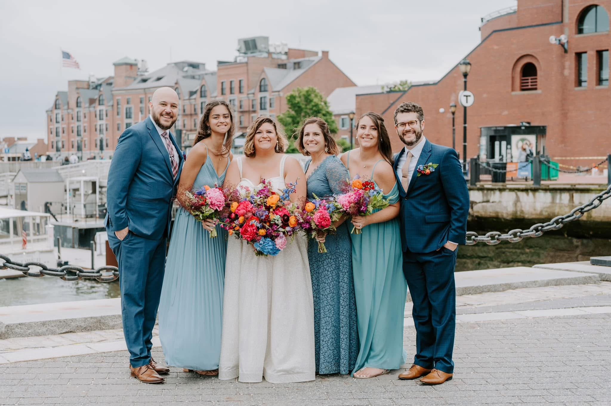 Group of six people dressed in formal attire, standing outdoors near a waterway and brick buildings, smiling and holding colorful bouquets of flowers.