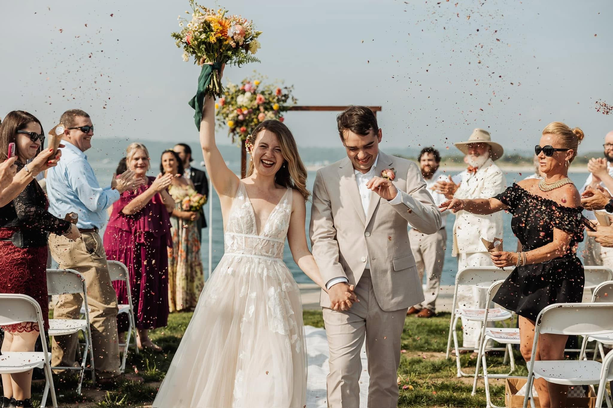 A newly married couple walking hand in hand through a celebration on the beach, with guests throwing rice and celebrating.