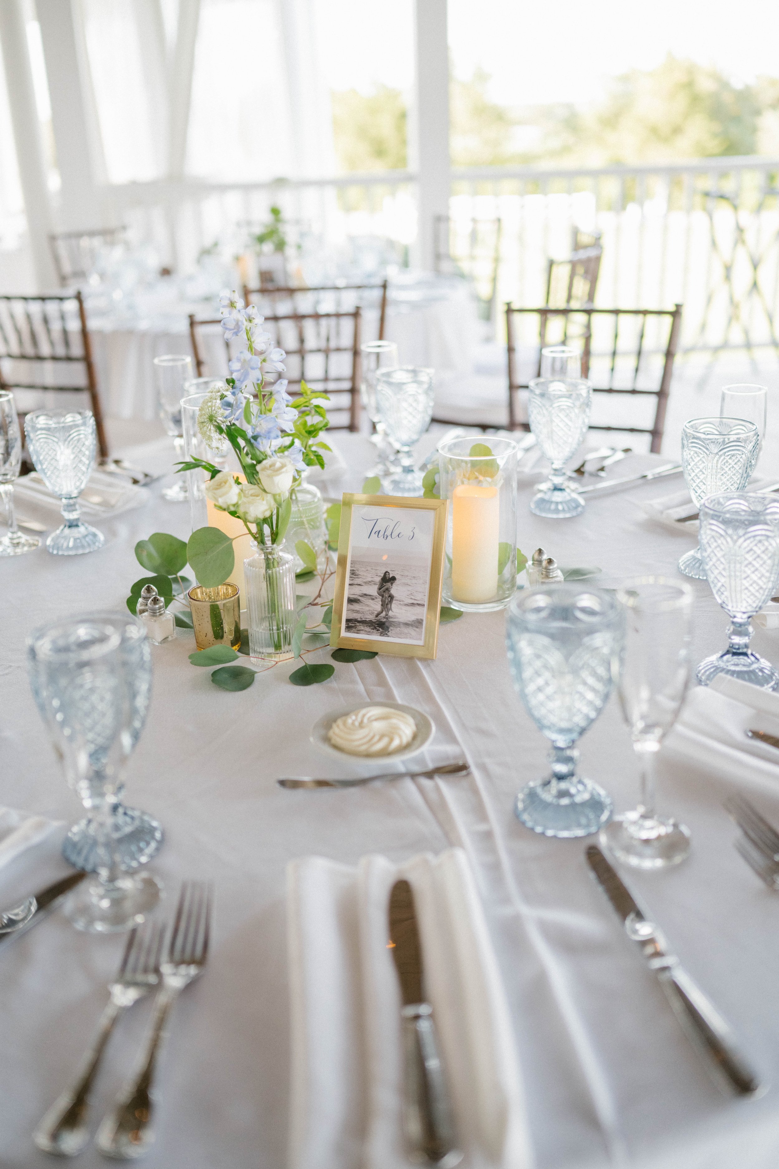 Upscale wedding reception table with white tablecloth, blue glassware, silver cutlery, floral centerpieces, candles, and framed photo card reading 'Table 3.'