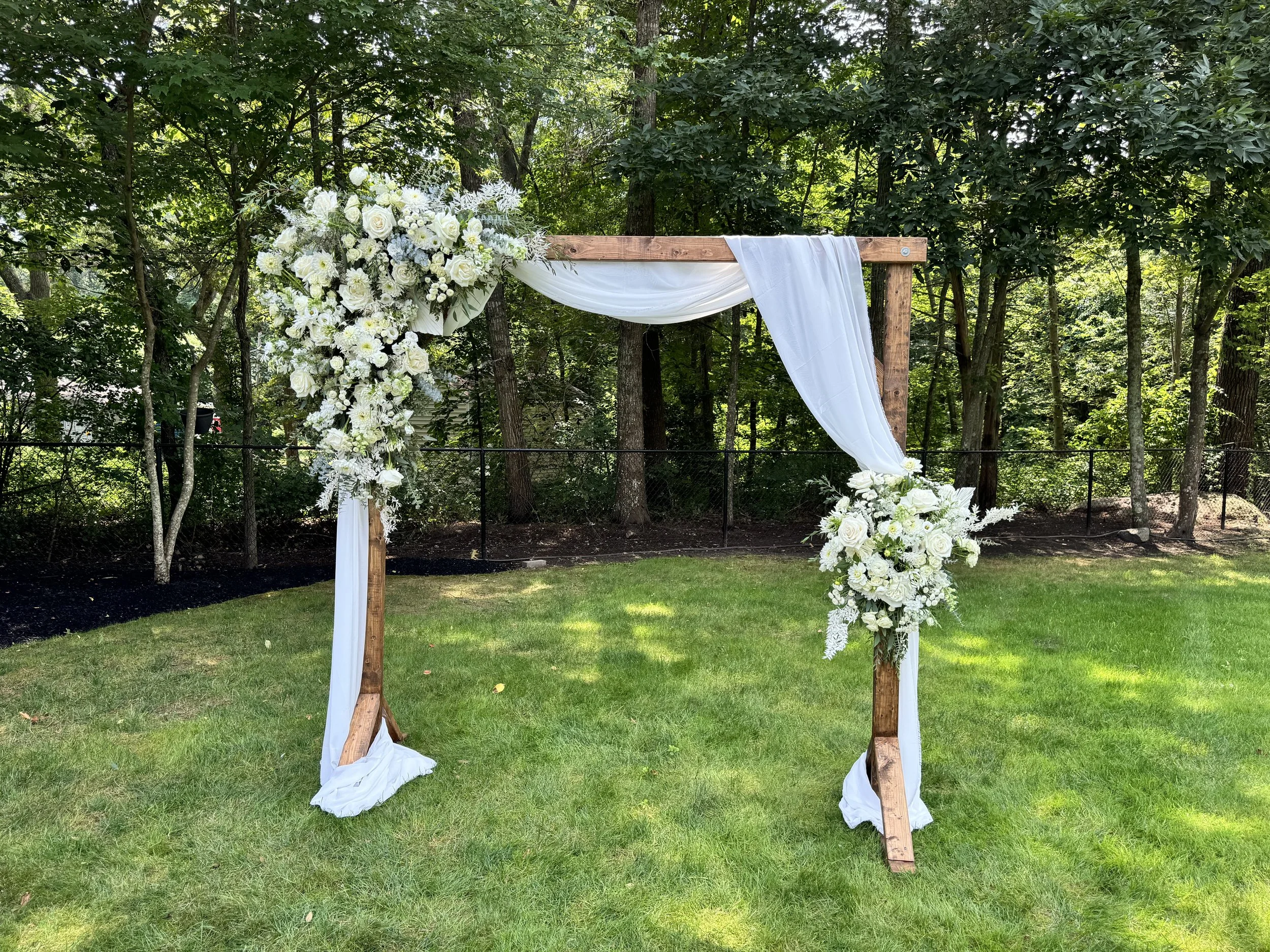 Wooden wedding arch decorated with white flowers and white drapery, set outdoors on grass surrounded by trees.