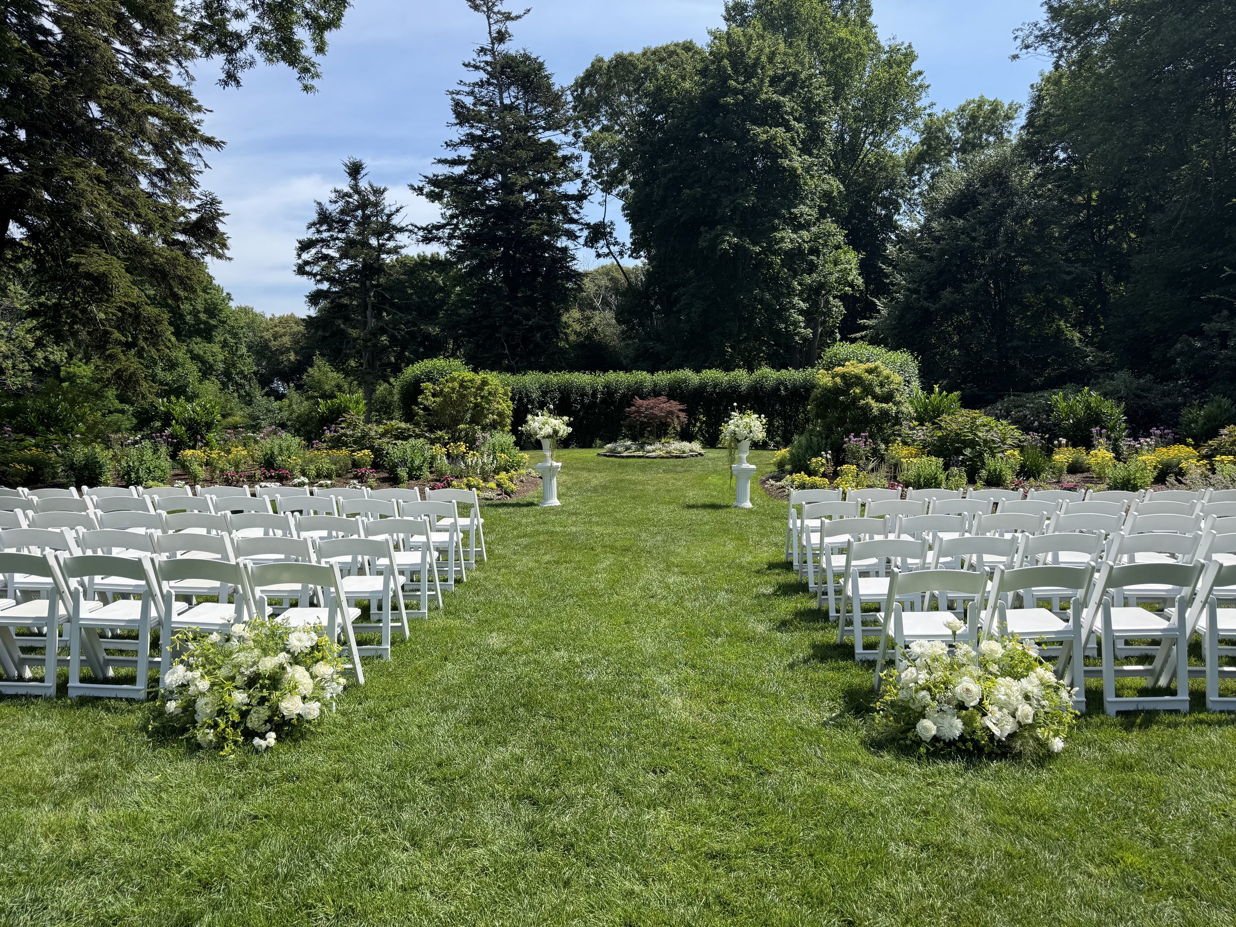 An outdoor wedding ceremony setup with aisle chairs and floral arrangements on a grassy lawn, surrounded by trees and colorful flowers.