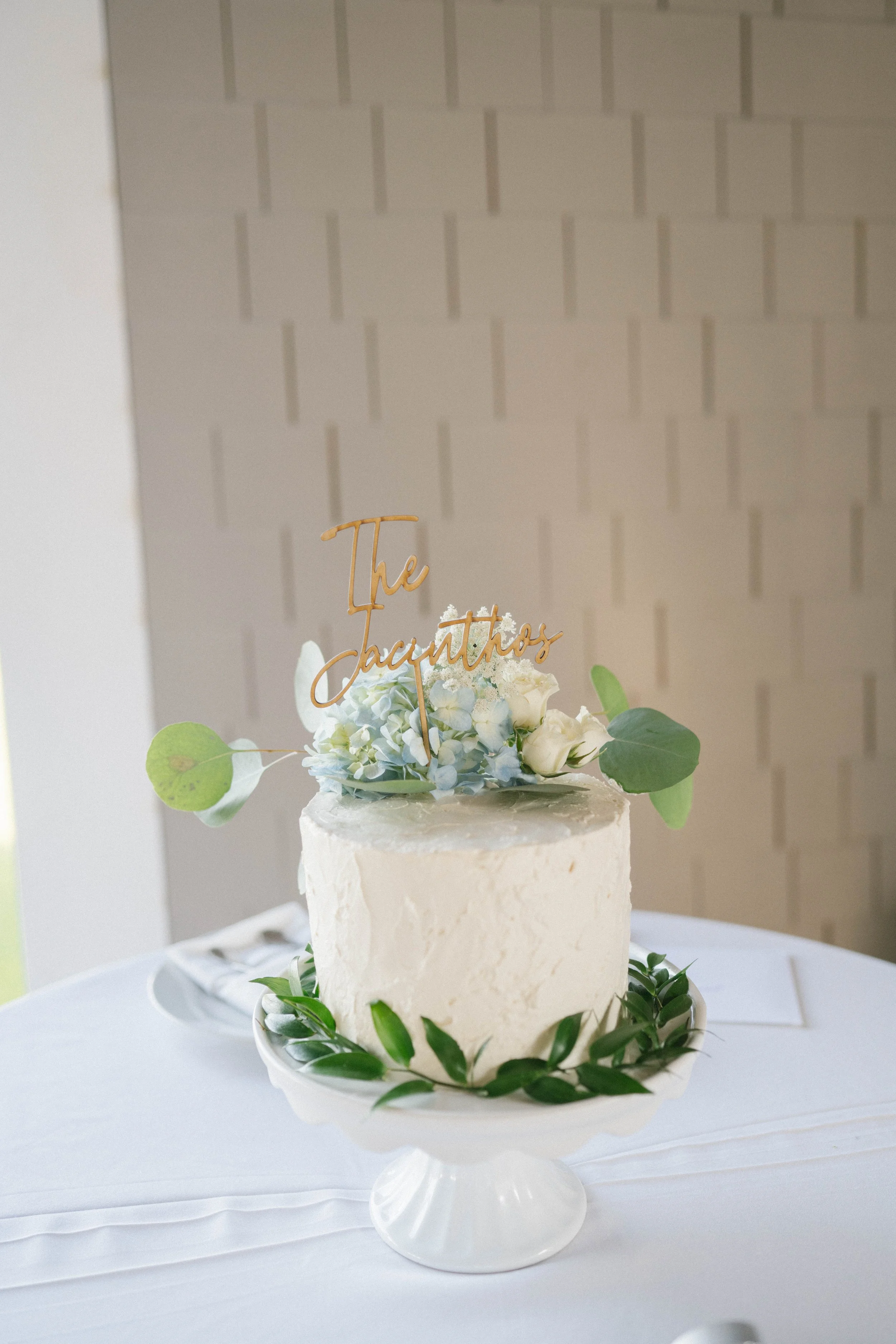 A white wedding cake with green leaves and white flowers on top, with a gold topper reading "The Jacintos," on a white cake stand on a table with a white tablecloth.