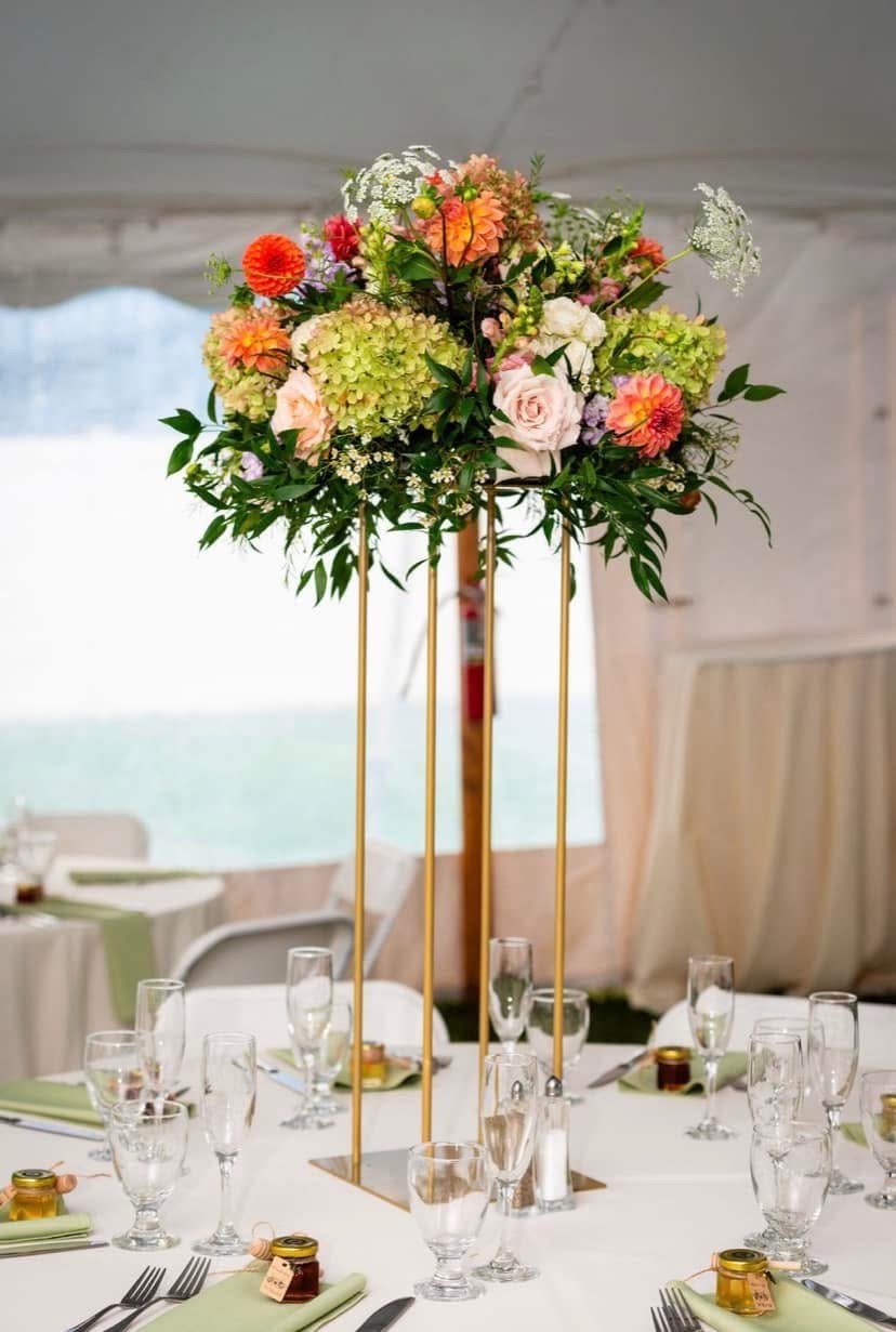 A tall floral centerpiece with various colorful flowers on a white table at an event, with glasses and table settings visible.
