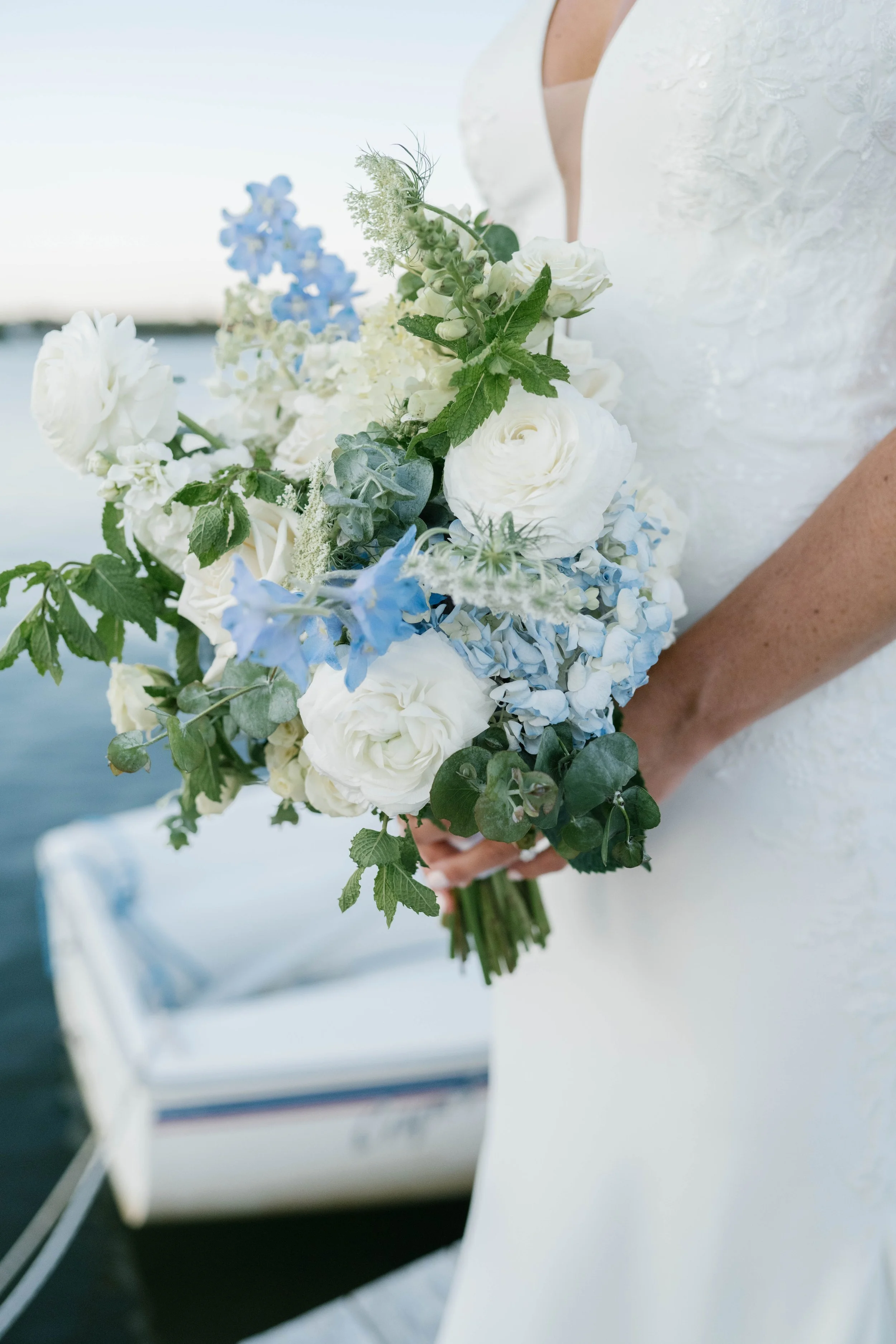 A bride holding a bouquet of white and light blue flowers near a body of water, with a boat in the background.
