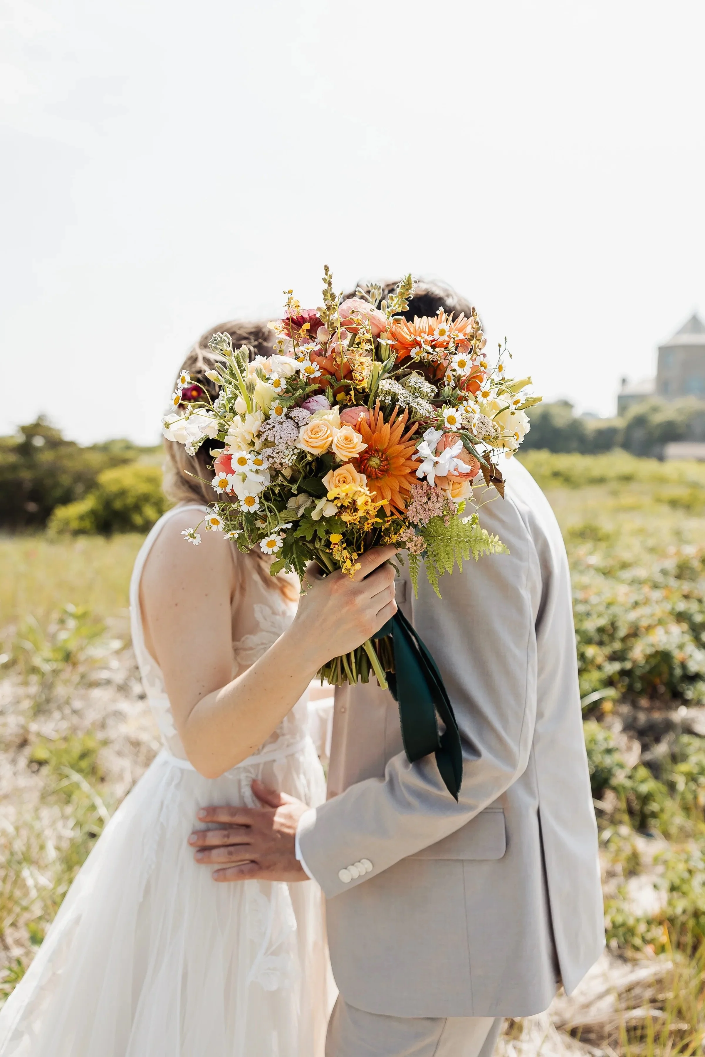 Couple kissing outdoors, hiding their faces behind a large bouquet of colorful flowers, with a bright sky and greenery in the background.