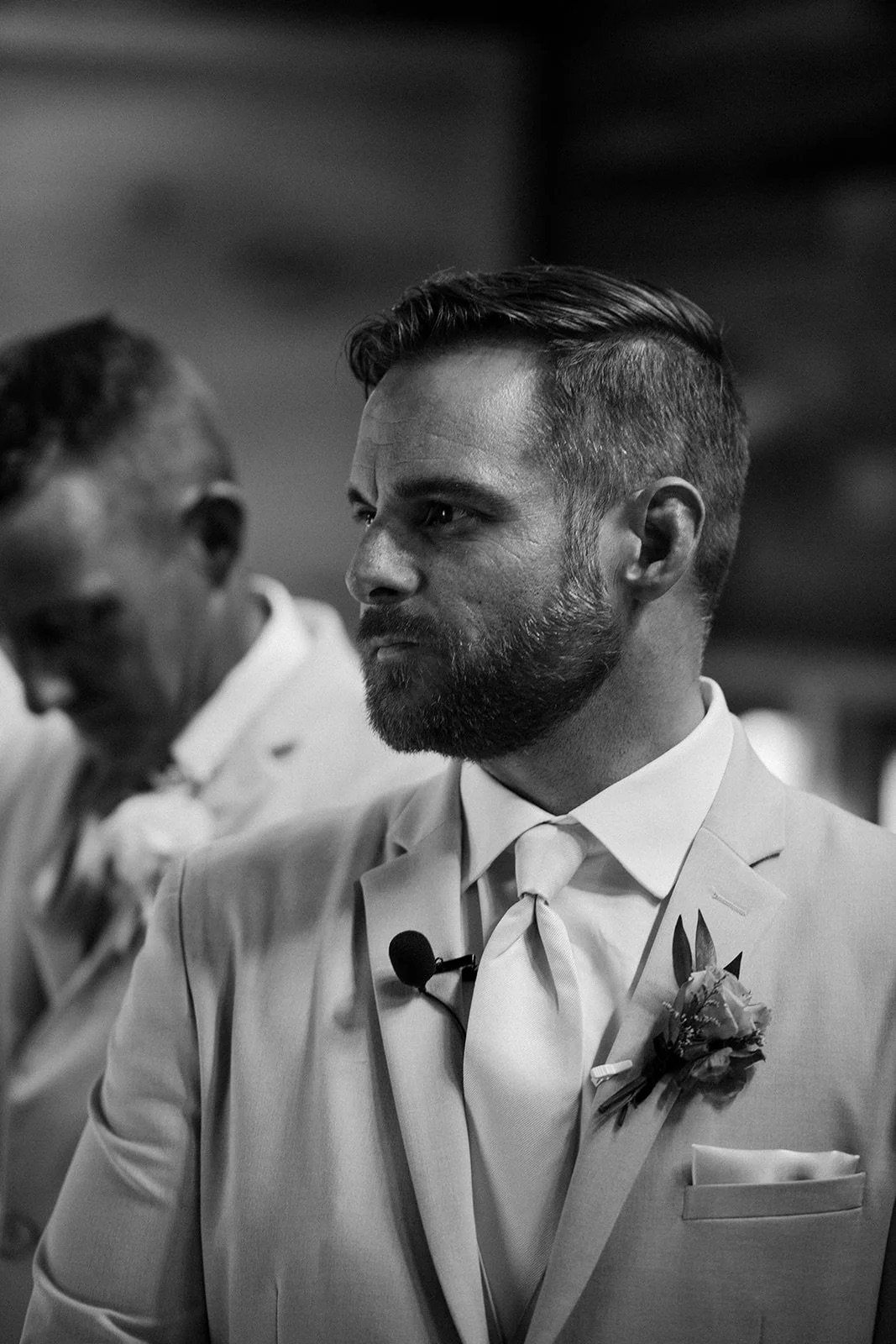 Black and white photo of a man in a suit with a boutonnière, wearing a tie clip, and a audio microphone clipped to his lapel, at a formal event. Another man is visible in the background.