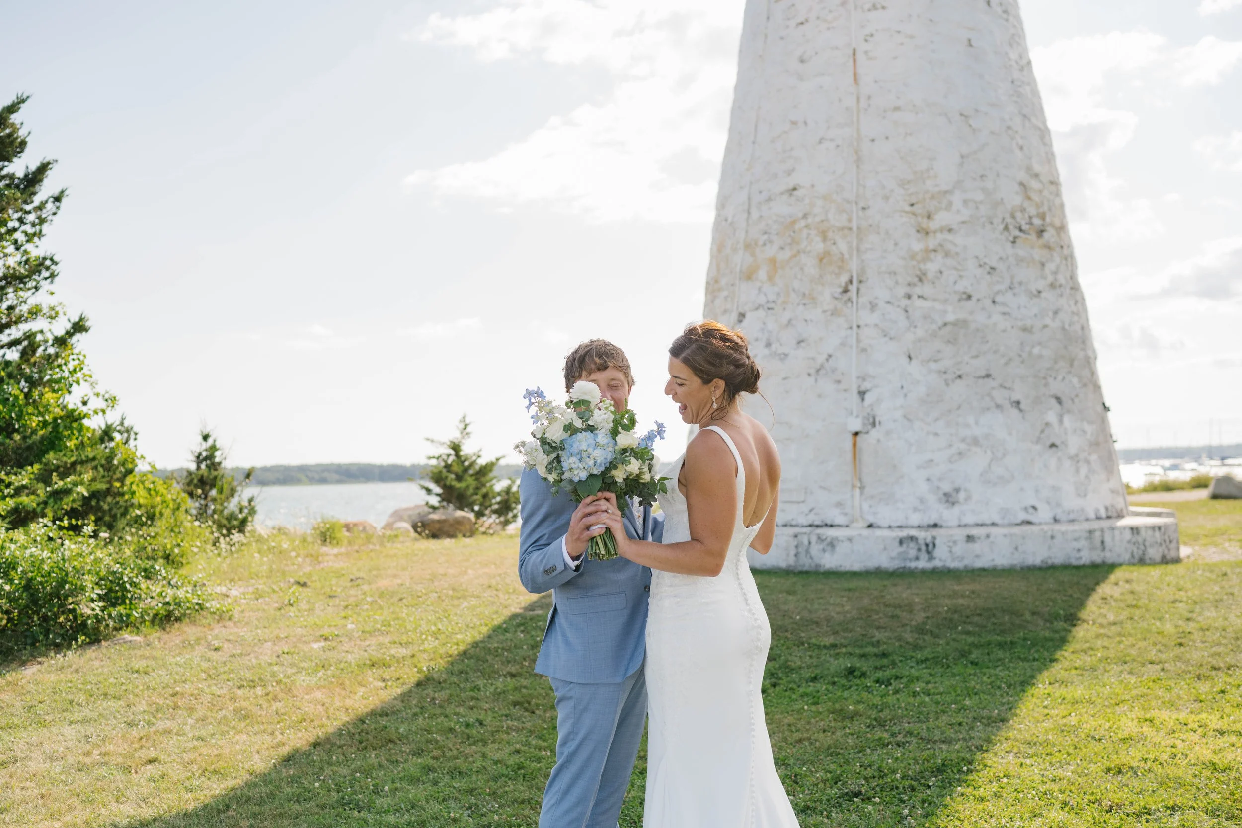 A bride and groom stand outdoors near a white lighthouse, with the groom holding a bouquet, as they smile and look at each other.
