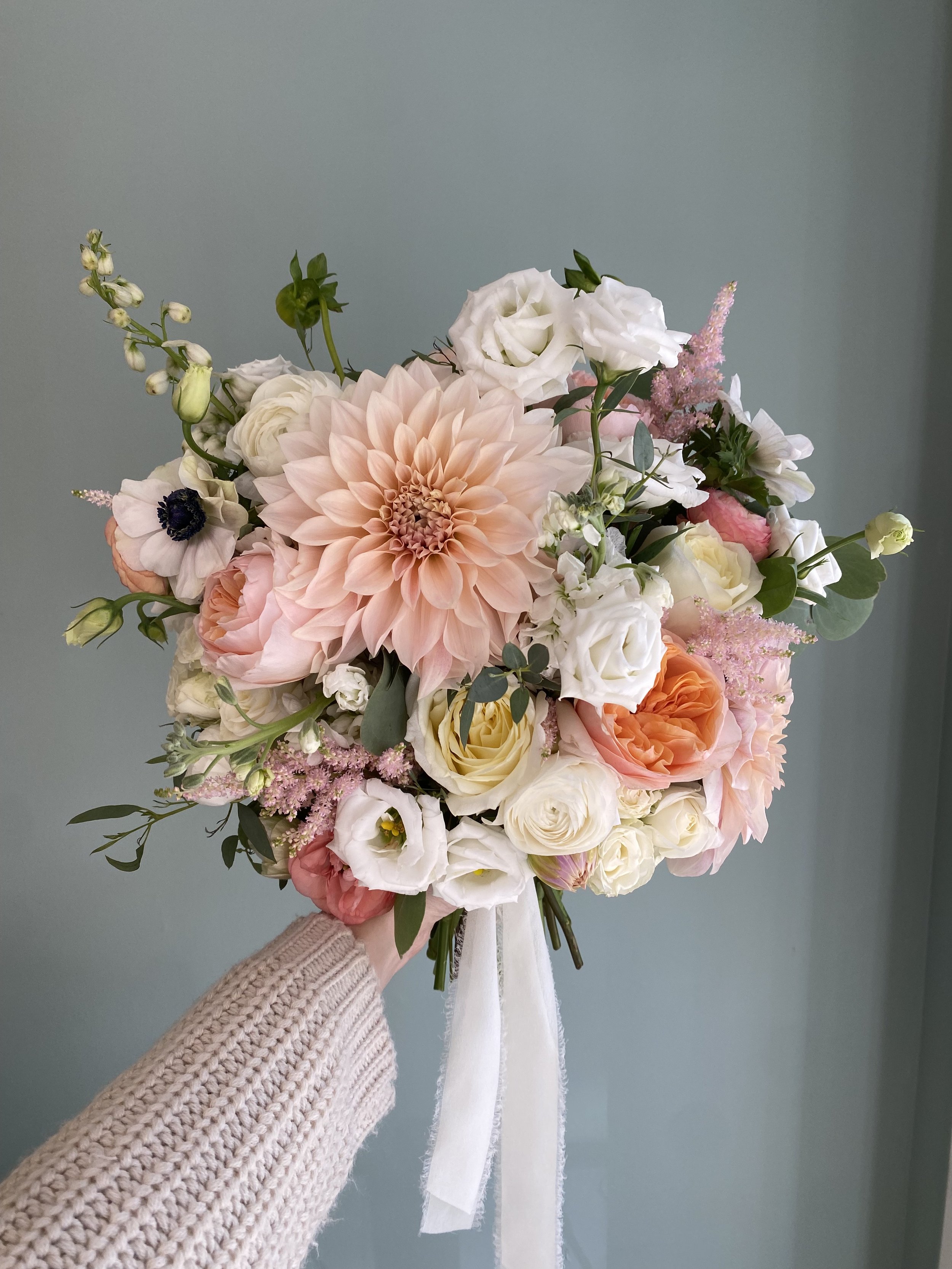 A bouquet of pink, white, and peach flowers held by a person wearing a beige sweater against a plain wall.