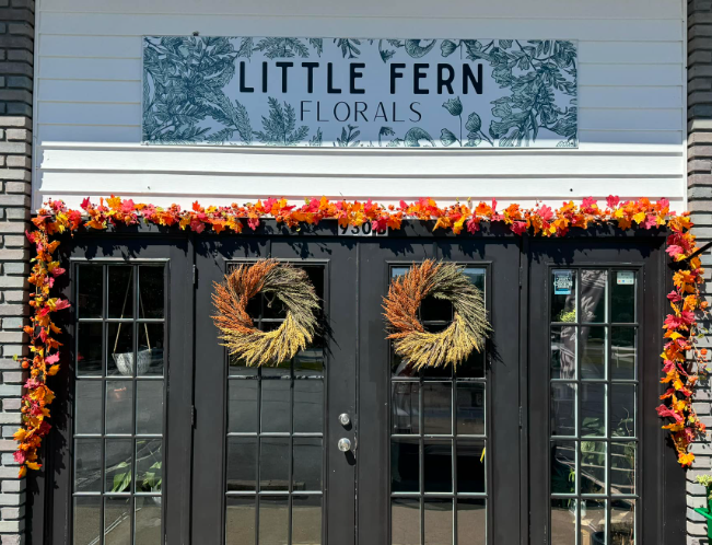 Exterior of a floral shop named Little Fern Florals with black double doors decorated with two wreaths and a floral garland framing the doors, and a sign above reading "Little Fern Florals" surrounded by leaf illustrations.