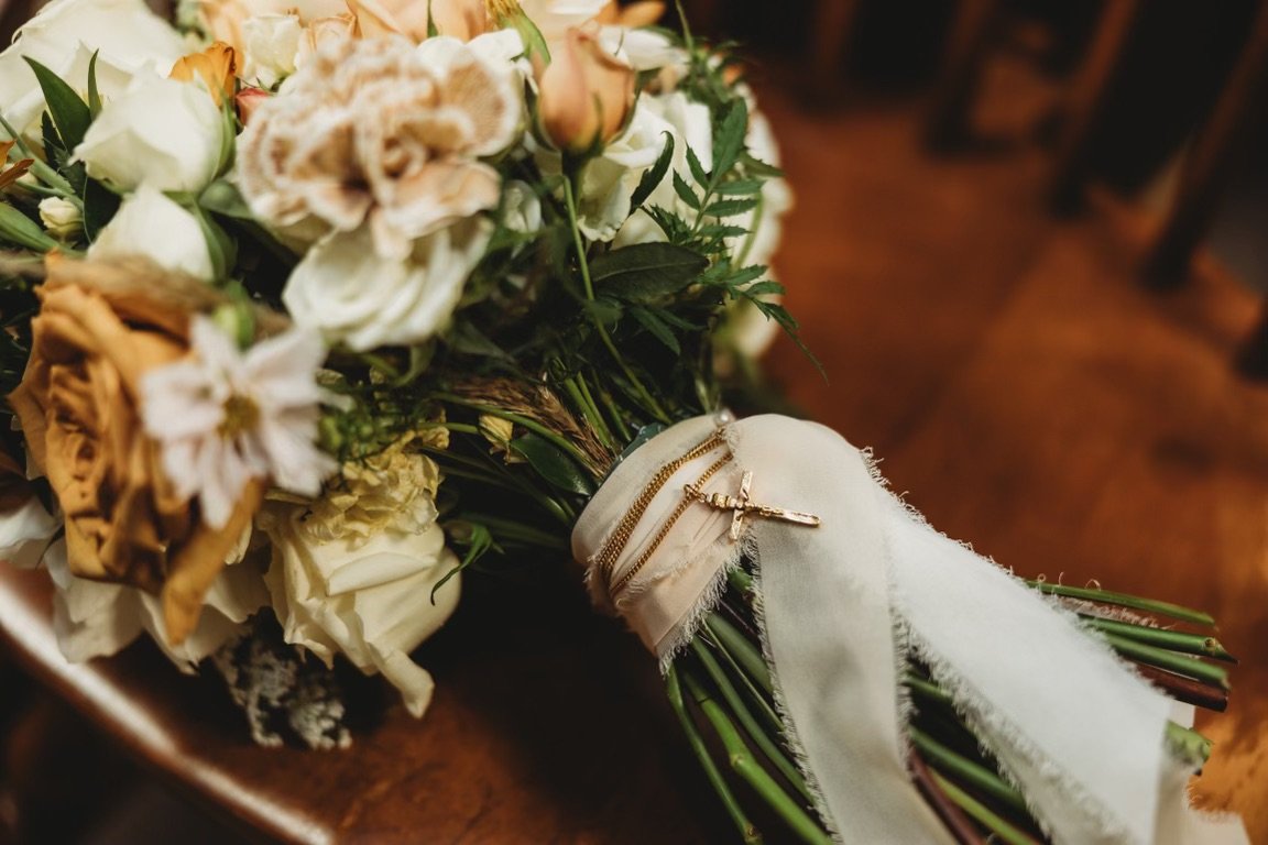 A bouquet of white, cream, and blush flowers with green foliage, tied with a white ribbon and decorated with jewelry, lying on a wooden surface.