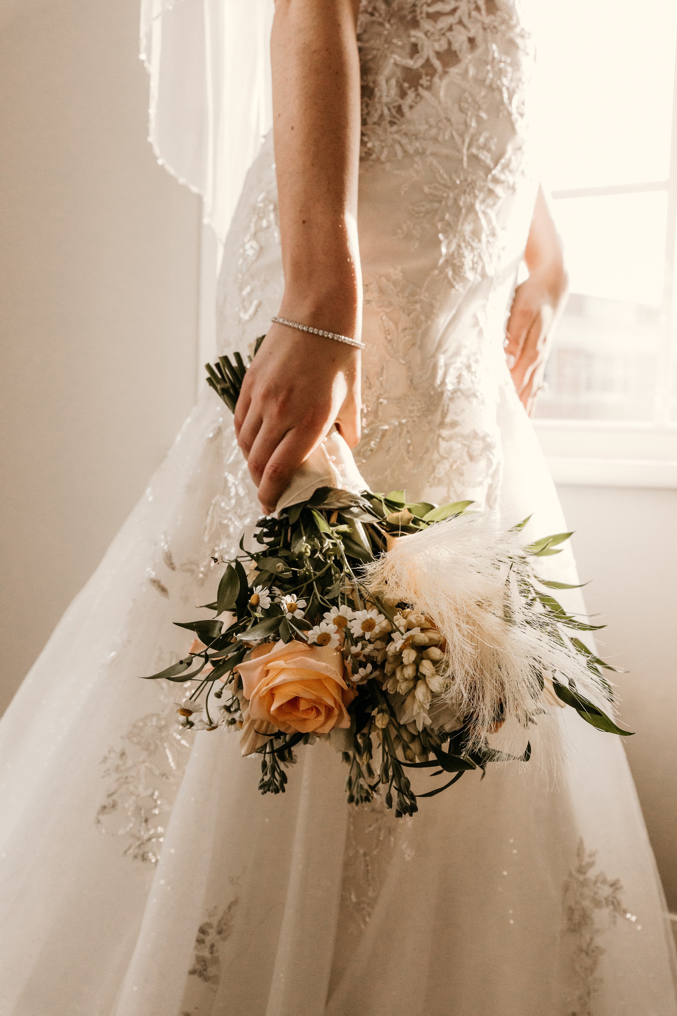 Close-up of a bride in an embroidered lace wedding dress holding a bouquet of peach roses, white daisies, and greenery in front of a window with sunlight.