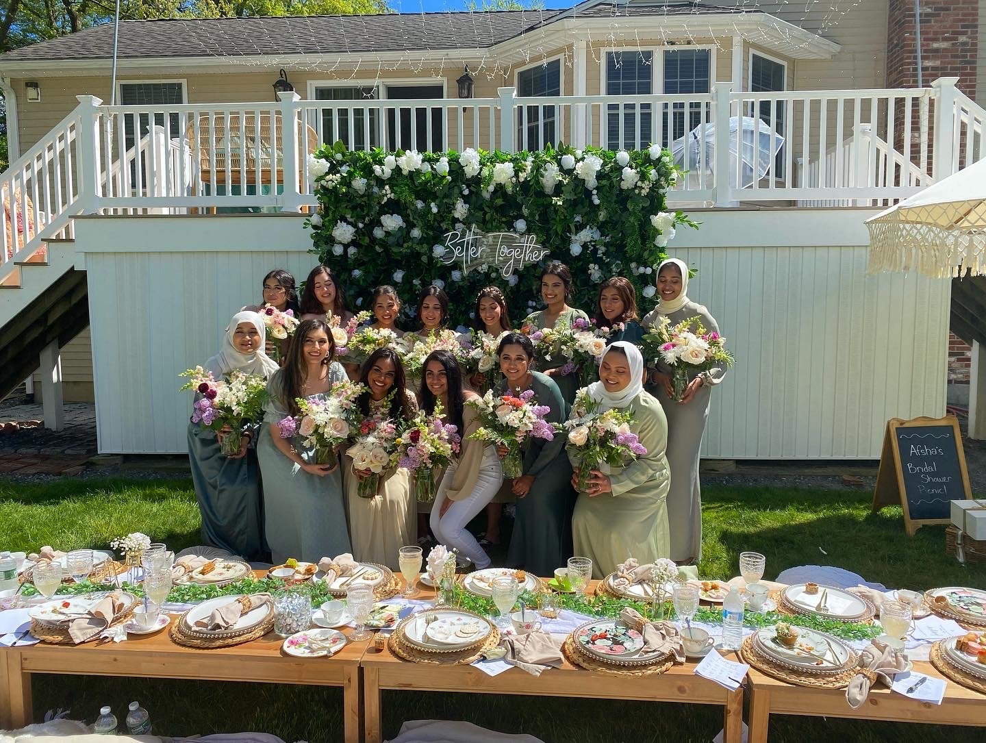 A group of women, some wearing hijabs, holding flower bouquets, gathered for a celebratory event outdoors on a sunny day. They are standing in front of a decorated backdrop with greenery and white flowers and a sign that reads 'Better Together.' A ta