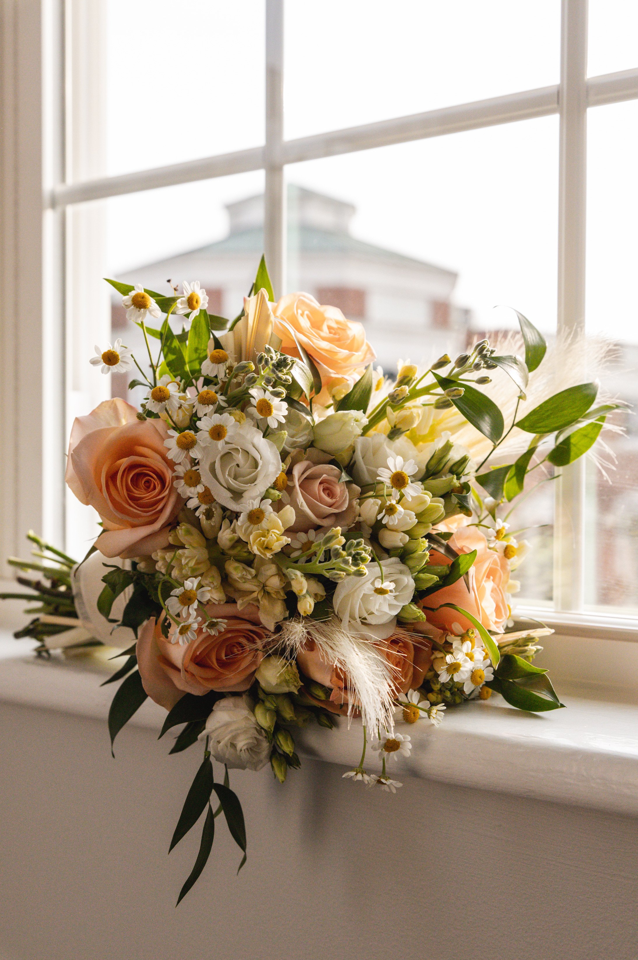 A bouquet of peach, white, and cream roses, daisies, and greenery resting on a windowsill near a bright window.