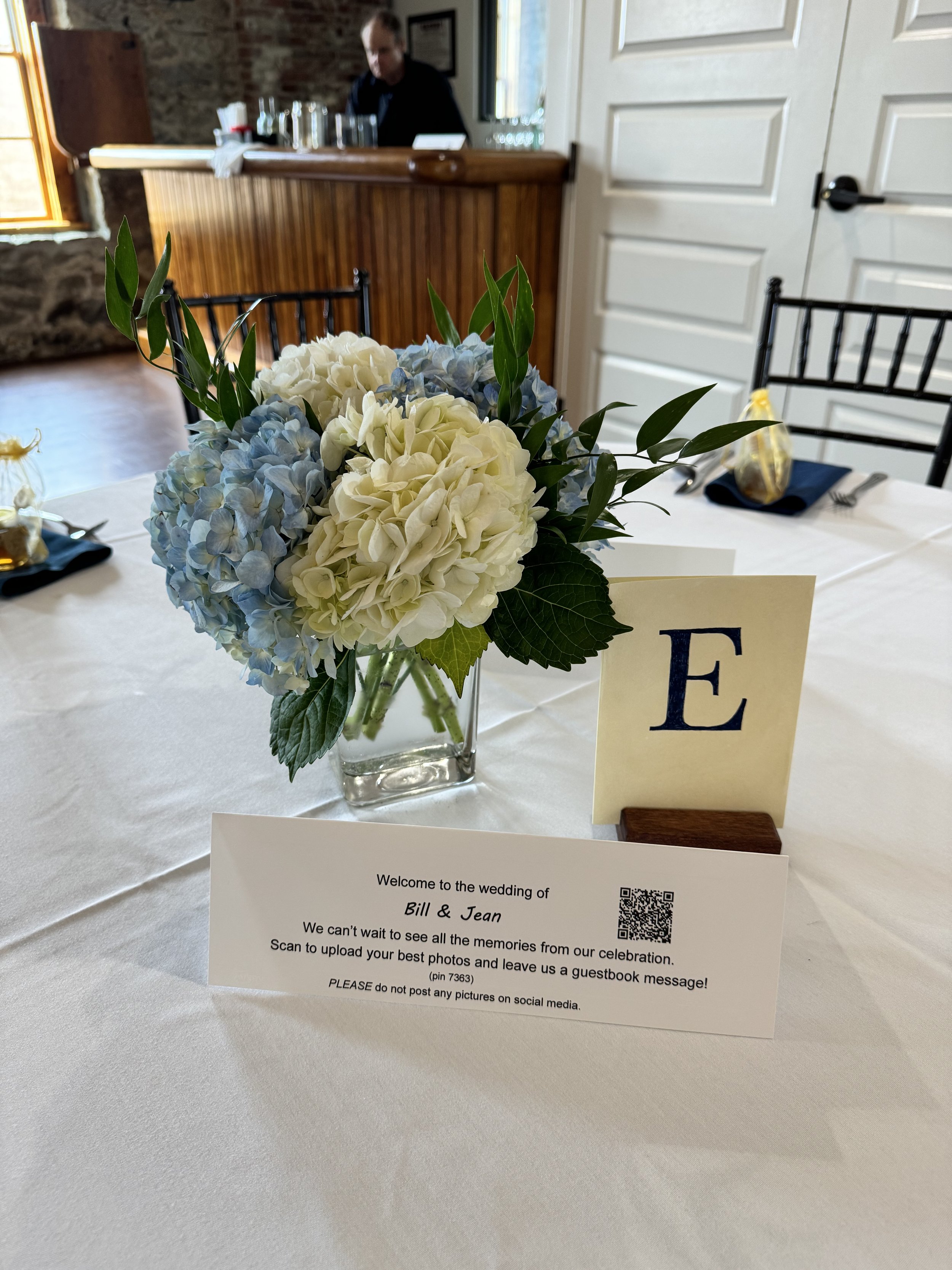 A wedding reception table with a small floral arrangement of white and blue hydrangeas in a glass vase, a table number card with a large letter E, and a welcome note for the wedding of Bill and Jean. In the background, a person is seen behind a woode