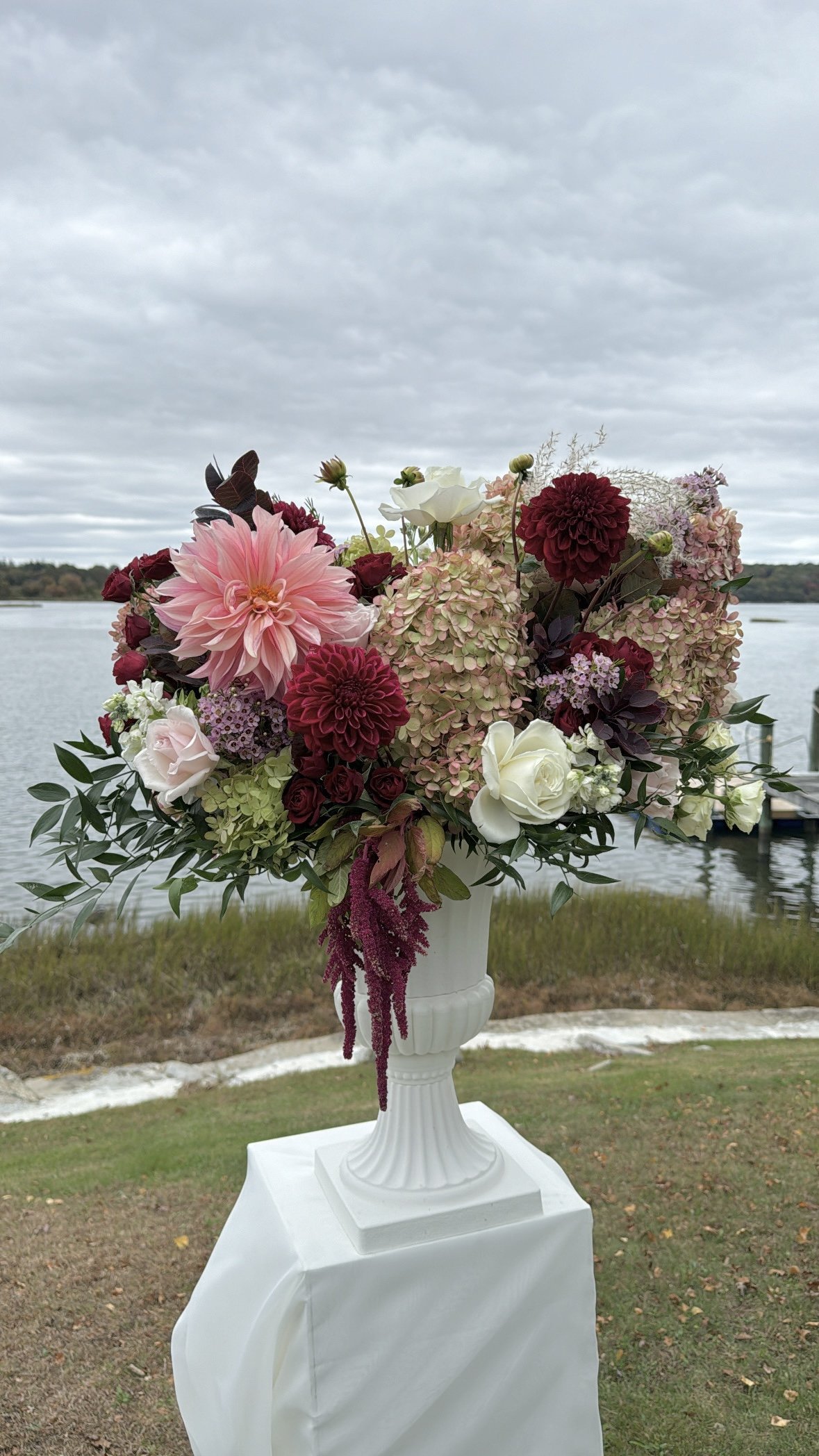 A large floral arrangement with pink dahlias, white roses, burgundy and pink hydrangeas, and other assorted flowers on a white pedestal near a body of water under a cloudy sky.