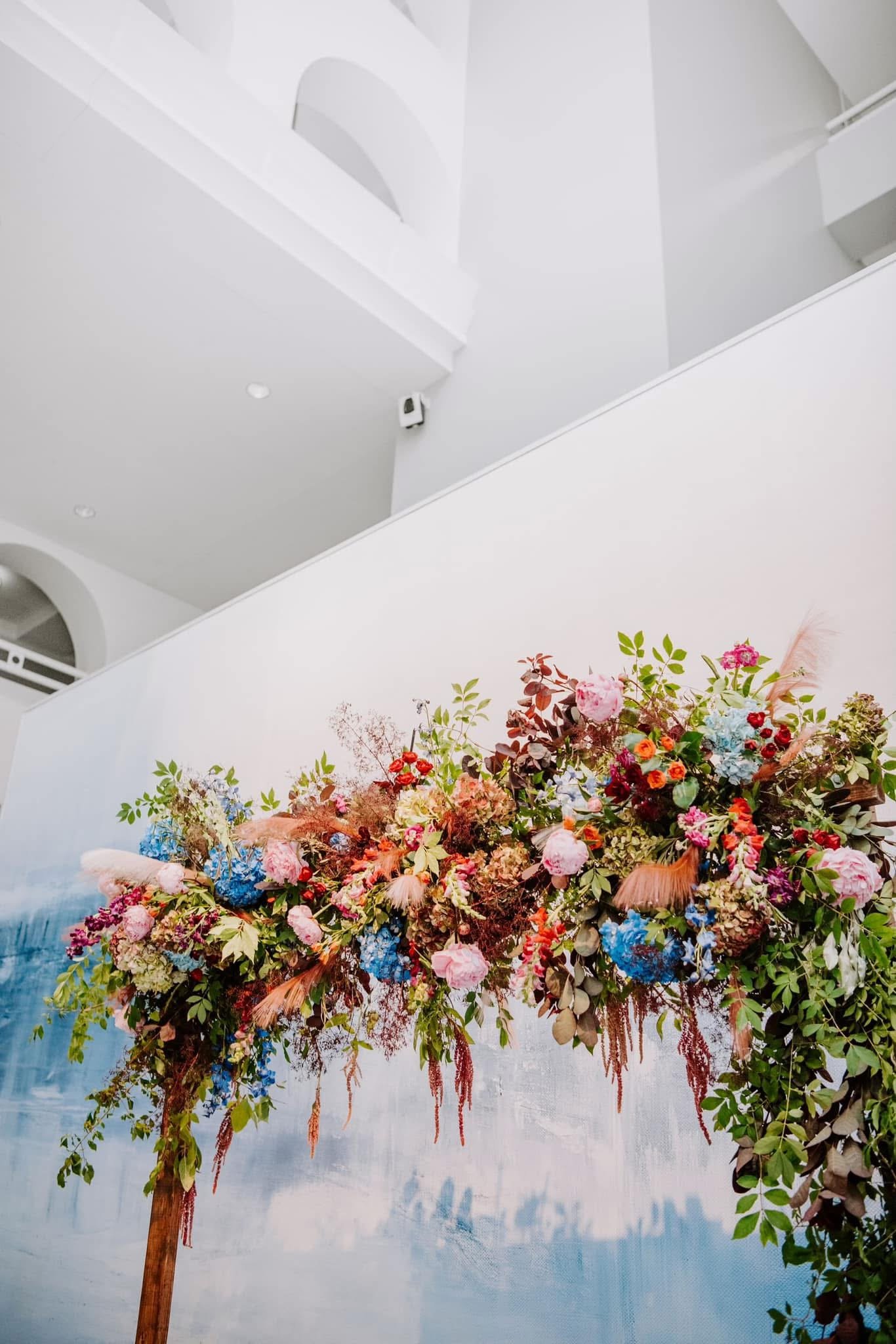 Colorful floral arrangement with pink, blue, and orange flowers and green foliage against a blue sky background.
