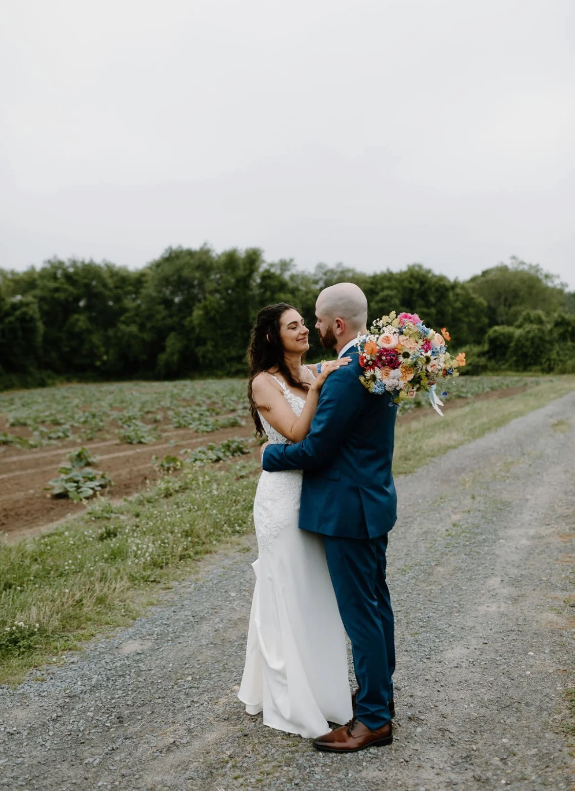 A bride and groom stand on a dirt pathway in a field, holding each other and looking into each other's eyes. The bride wears a white wedding dress, and the groom wears a blue suit. The groom holds a large, colorful bouquet of flowers. Trees and a clo
