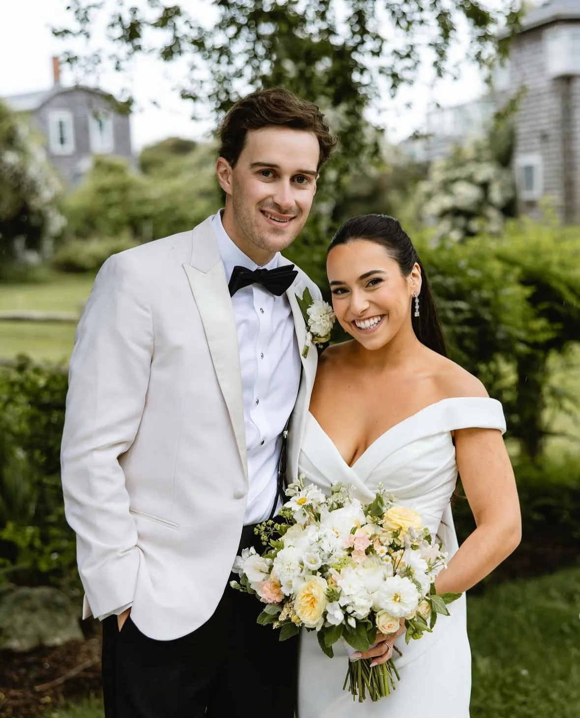 A smiling bride and groom pose outdoors on their wedding day, with the groom in a white tuxedo jacket and the bride in a white off-the-shoulder wedding dress, holding a bouquet of white and pale pink flowers.